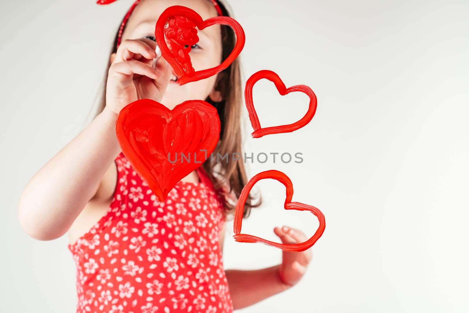 Little girl with long curly hair in red clothes draw hearts with paint on transparent glass smiling happily on white background. Family, love, holidays, birthday, Mother's day concept. Copy space.