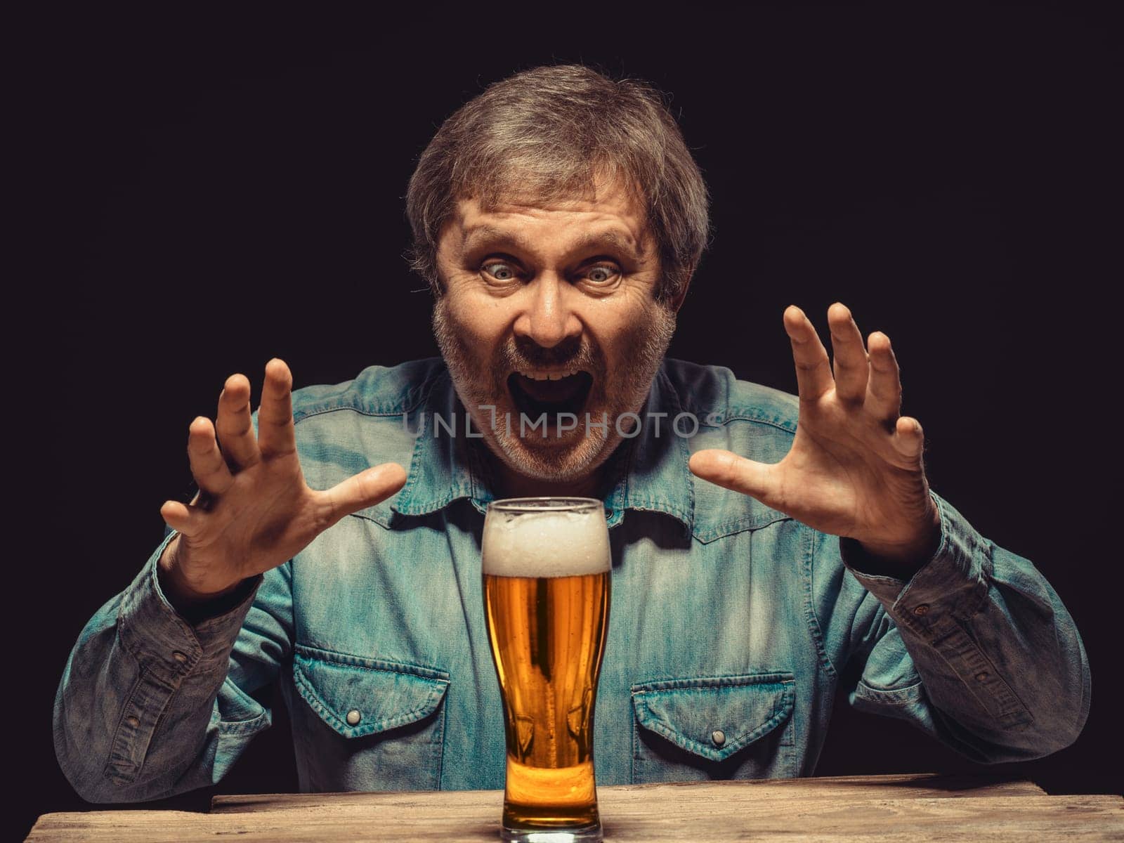 Enjoying his favorite beer.  The front view of handsome screaming man as fan in denim shirt with glass of beer, sitting at the wooden table. Concept of enthusiasm and ecstasy