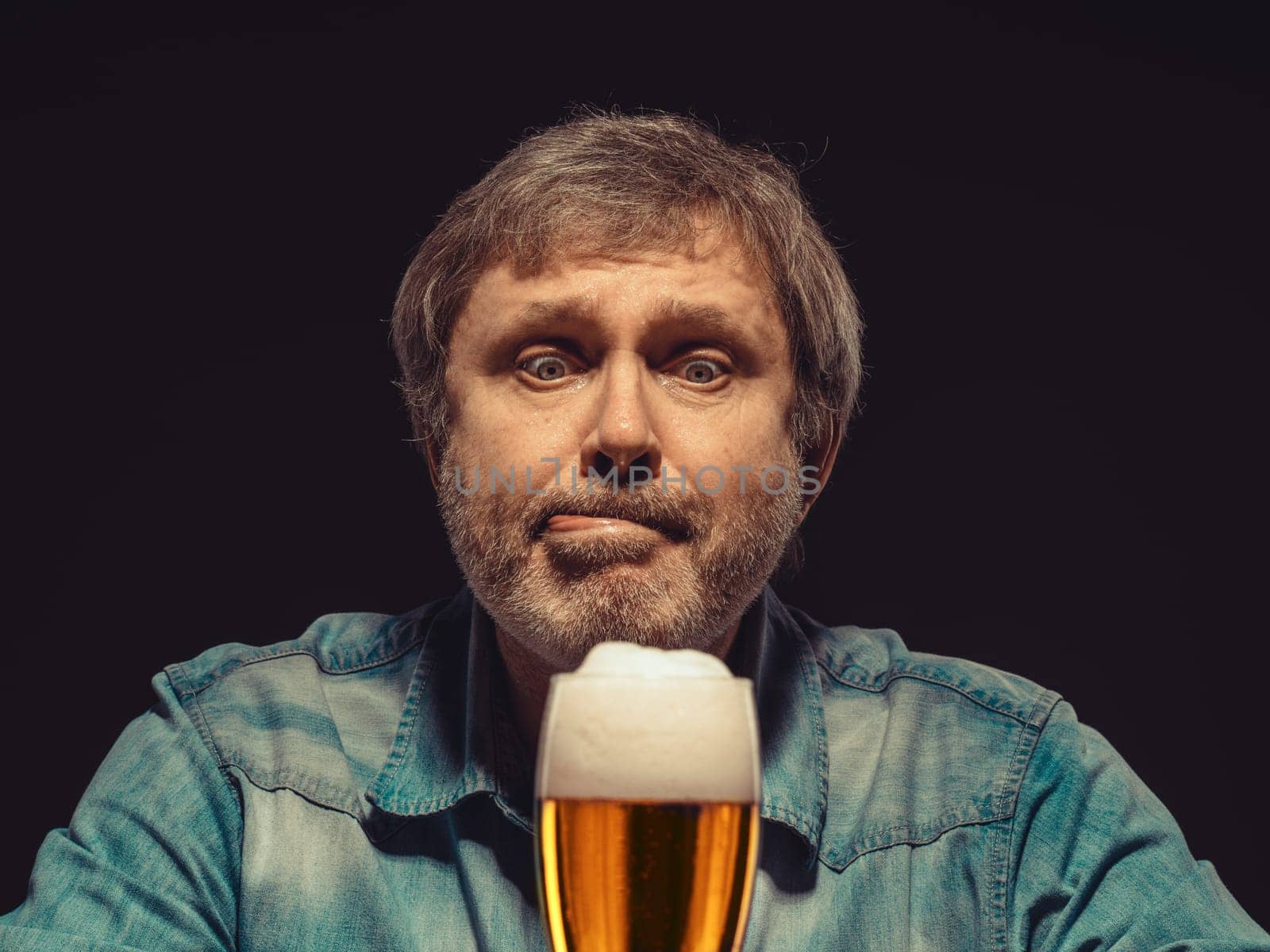 Enjoying his favorite beer.  The front view of handsome spellbound man in denim shirt with glass of beer, sitting at the wooden table. Concept of desire and love for beer