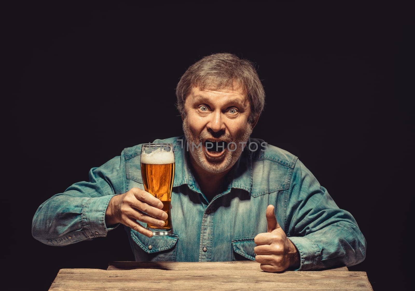Enjoying his favorite beer.  The front view of handsome smiling  man as fan in denim shirt with glass of beer, sitting at the wooden table