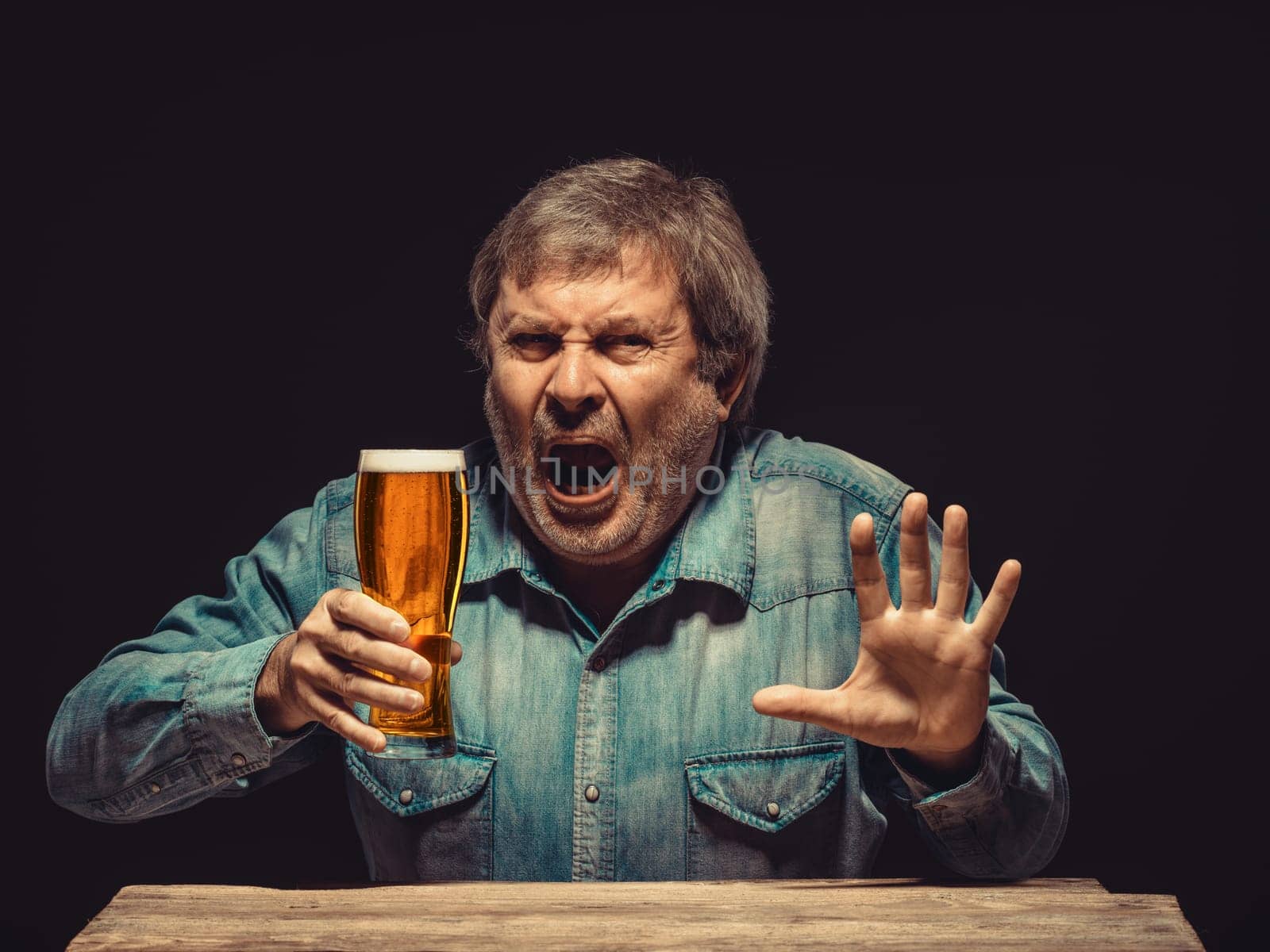 The front view of handsome man as emotional fan in denim shirt with glass of beer, sitting at the wooden table. Concept of fierce fan
