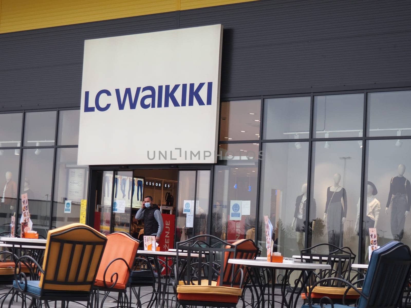 Sremska Mitrovica, Serbia, February 27, 2021 Shopping center. Clothing store LC WAIKIKI. A man wearing a surgical protective mask walks out of a shop's sliding doors. Cafe with metal chairs and tables