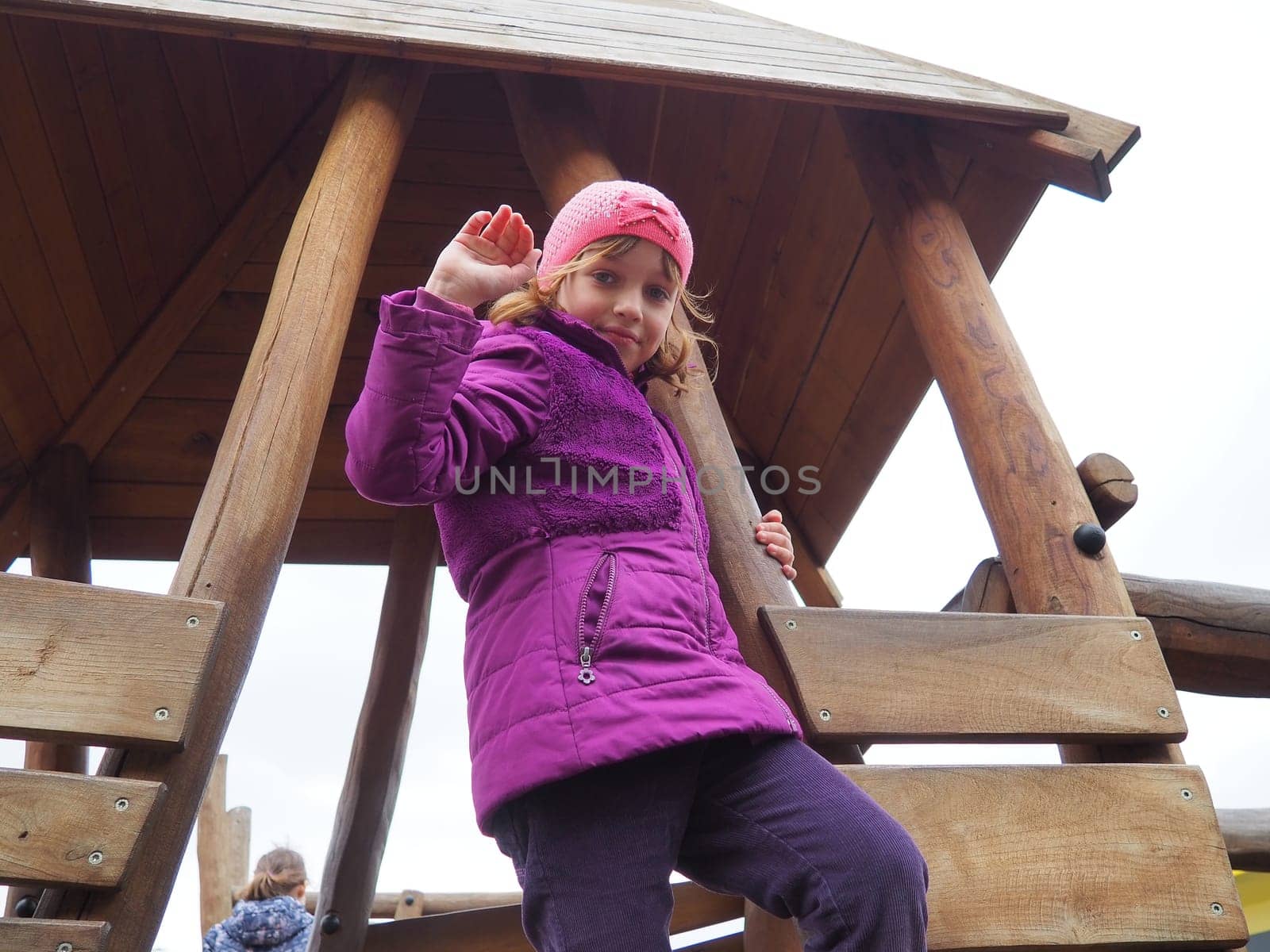 girl 7 years old on the playground. The child is dressed in warm demi-season clothes - a purple jacket and a pink hat. Wooden playground for climbing. Girl waving right hand.
