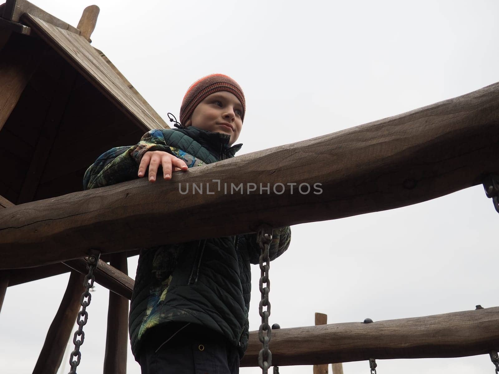 boy 8 years old on the playground. The child is dressed in warm, demi-season clothes - a brown hat and a green jacket. Children's wooden climbing area. Boy looking away.