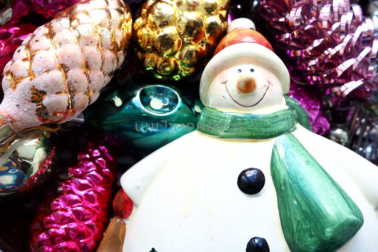 Cheerful smiling ceramic snowman in a green scarf and winter hat. New Year and Christmas decorations, tinsel. Multi-colored balls in the form of cones. Festive interior decor. New Year or Christmas.