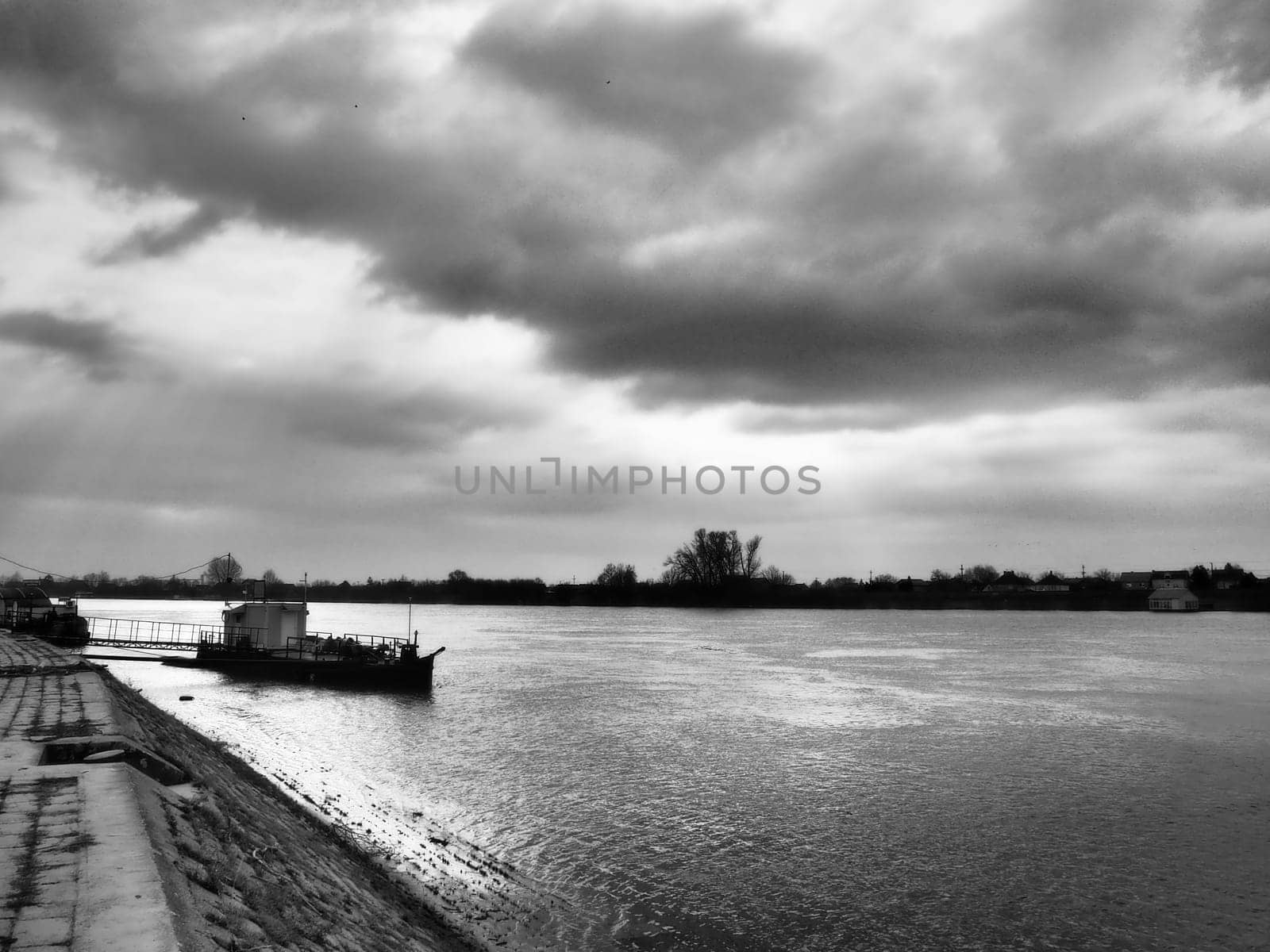 River landscape with a lonely ship at anchor. Cloudy weather with heavy clouds. Reflection of clouds in the water. The opposite bank is the horizon. Sava river, bank of Sremska Mitrovica, Serbia