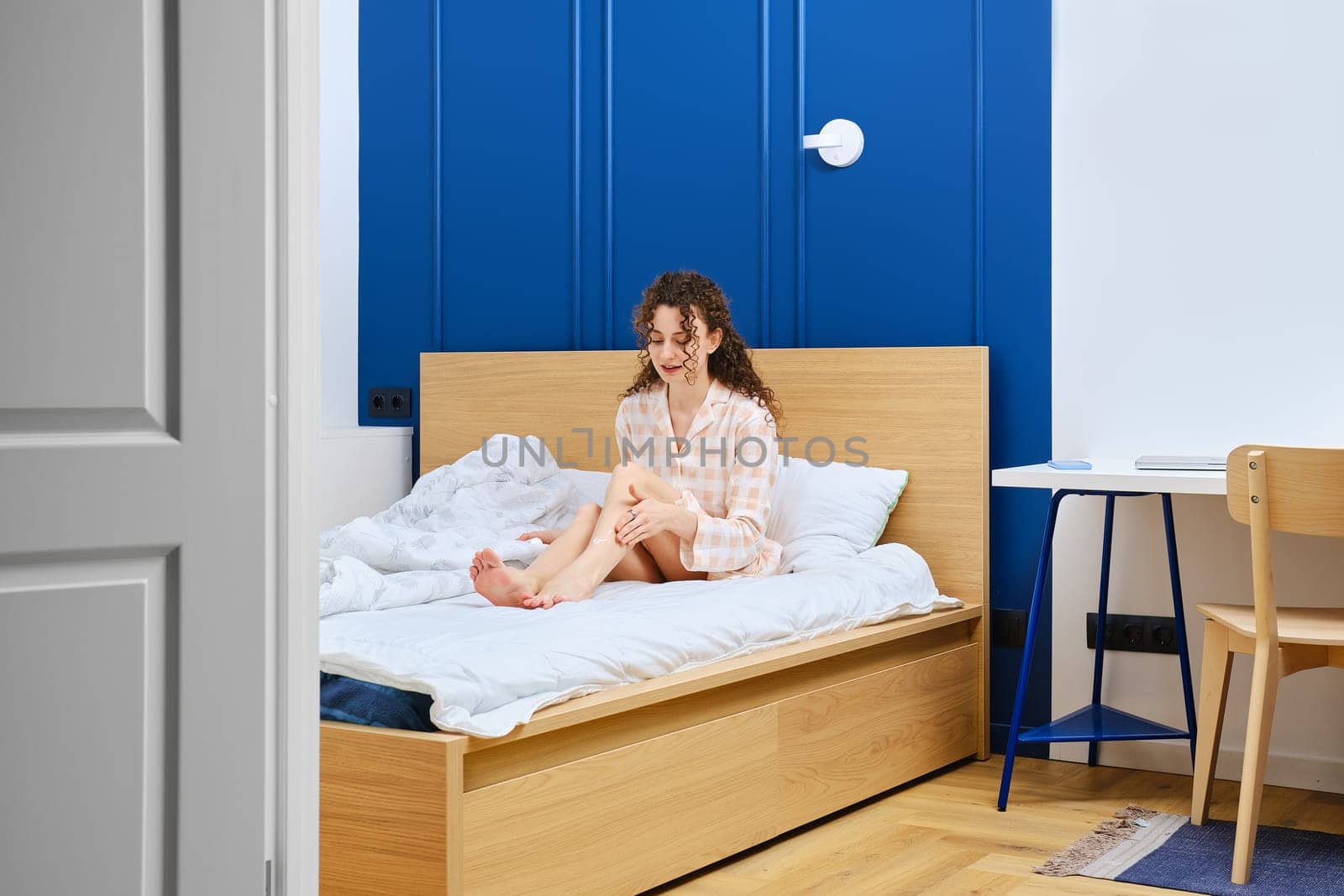 Young woman applies soothing aftershave cream to her feet sitting in bed in bathrobe
