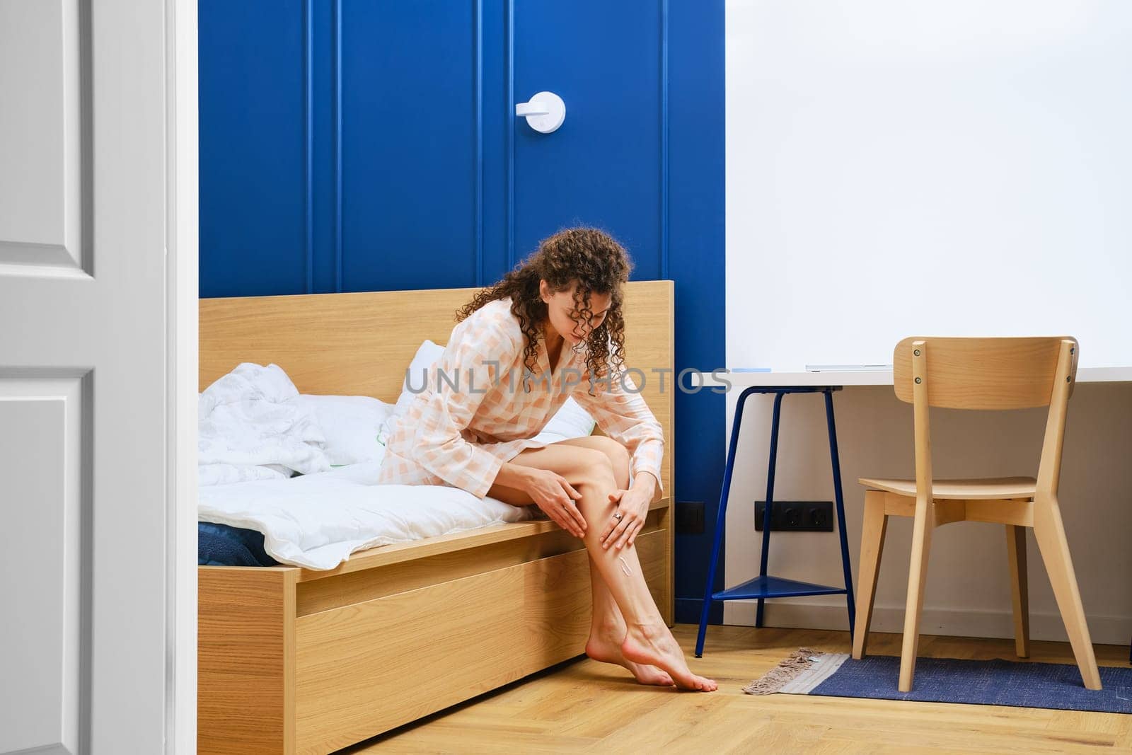 Young woman in bathrobe sitting in bedroom and rubbing cream on her legs