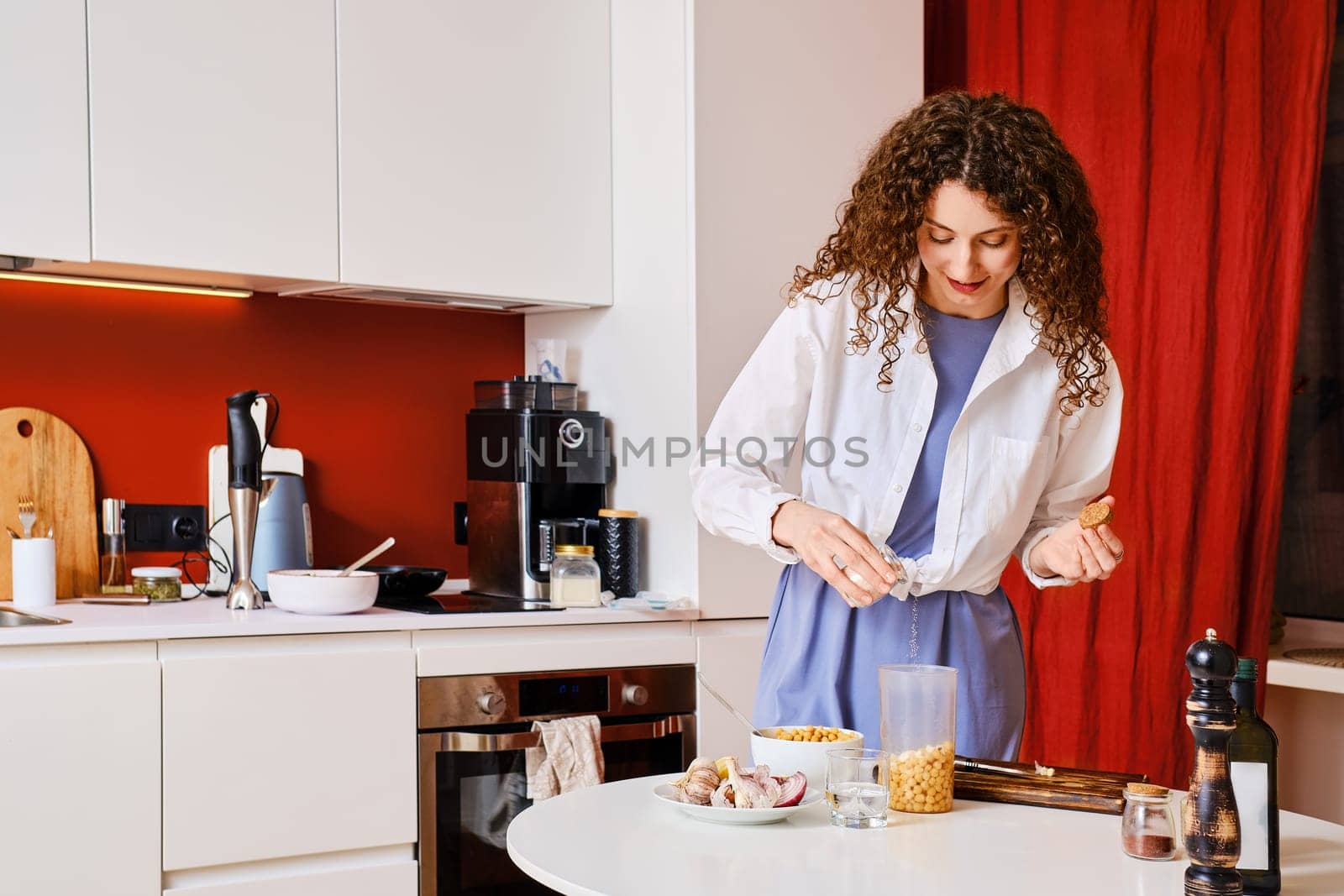 Young housewife adding salt into blender with chickpeas while making hummus