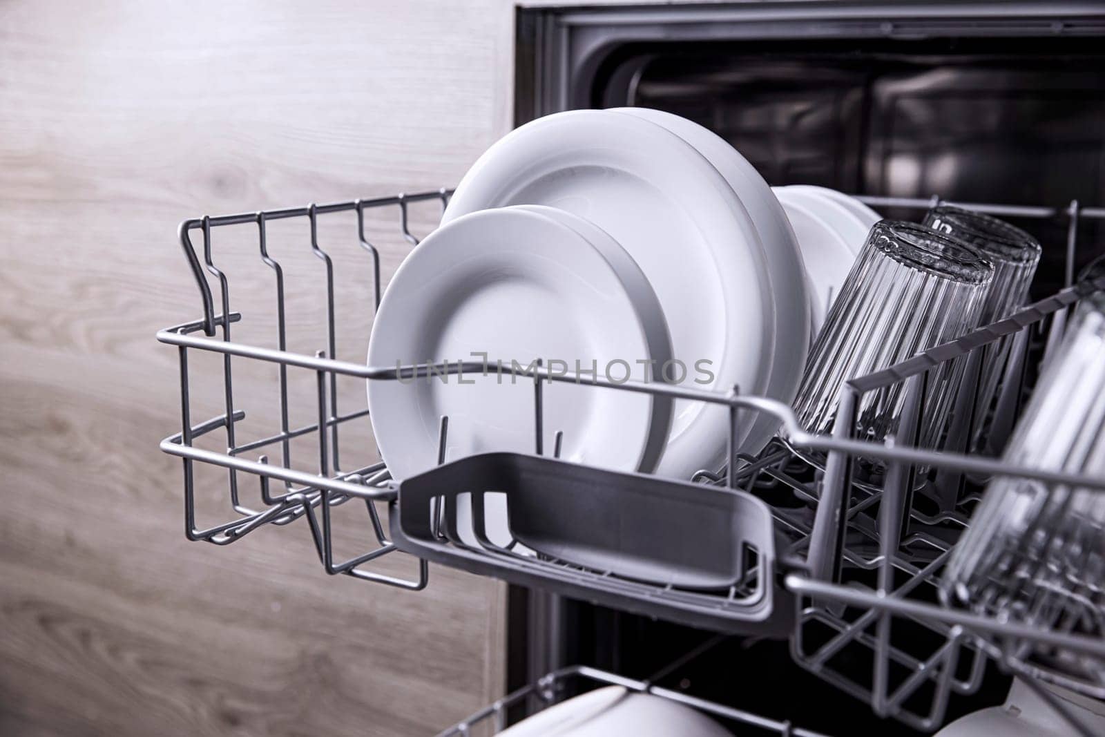 Front view of open automatic stainless steel built-in fully integrated top control dishwasher range machine with clean utensils, cutlery, glasses, dishes, plates inside on the upper deck kitchen