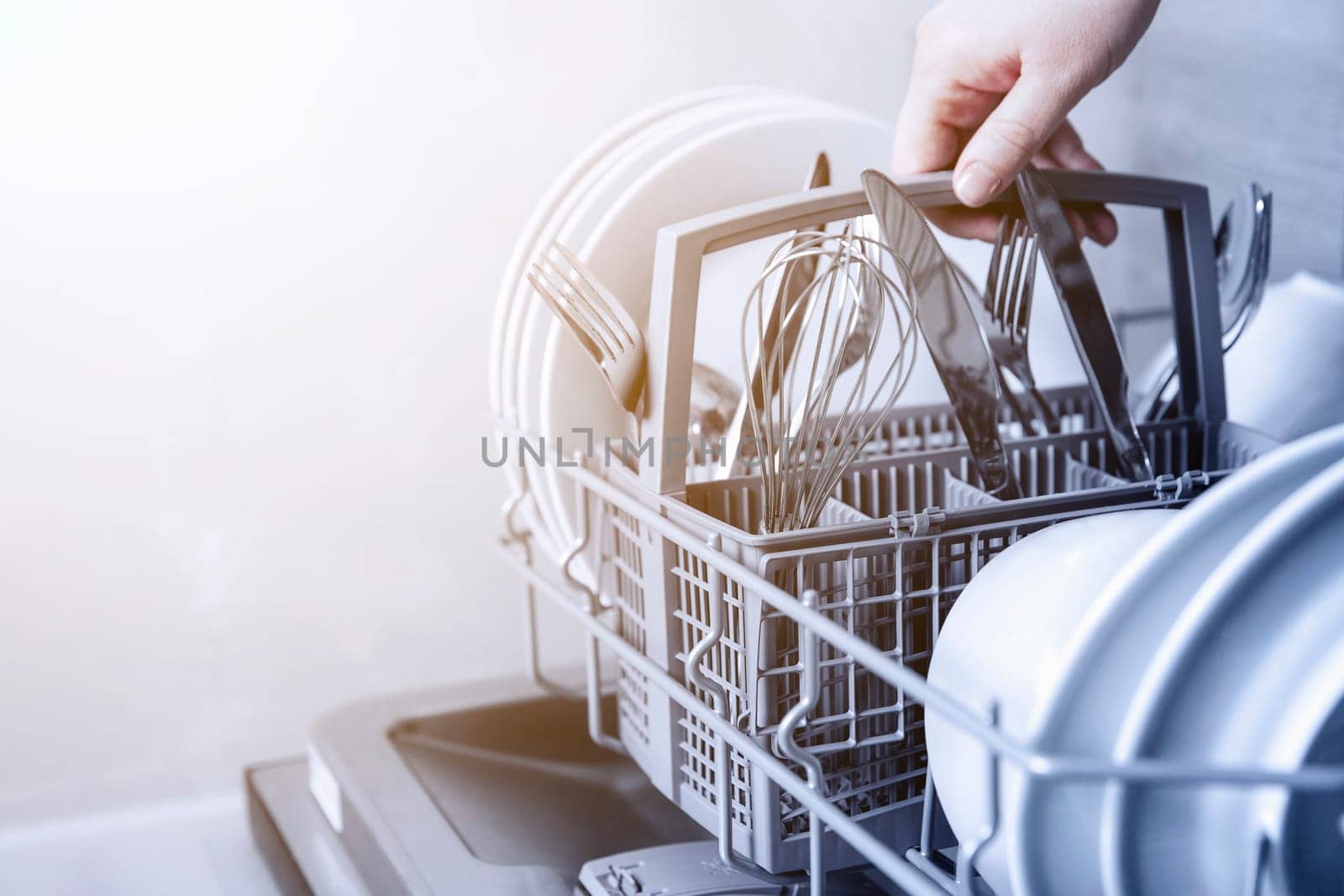 Close-up of female hand holding full cutlery basket with clean knife, fork, whisk. Loading to, empty out or unloading from open automatic dishwasher machine with clean utensils in home kitchen