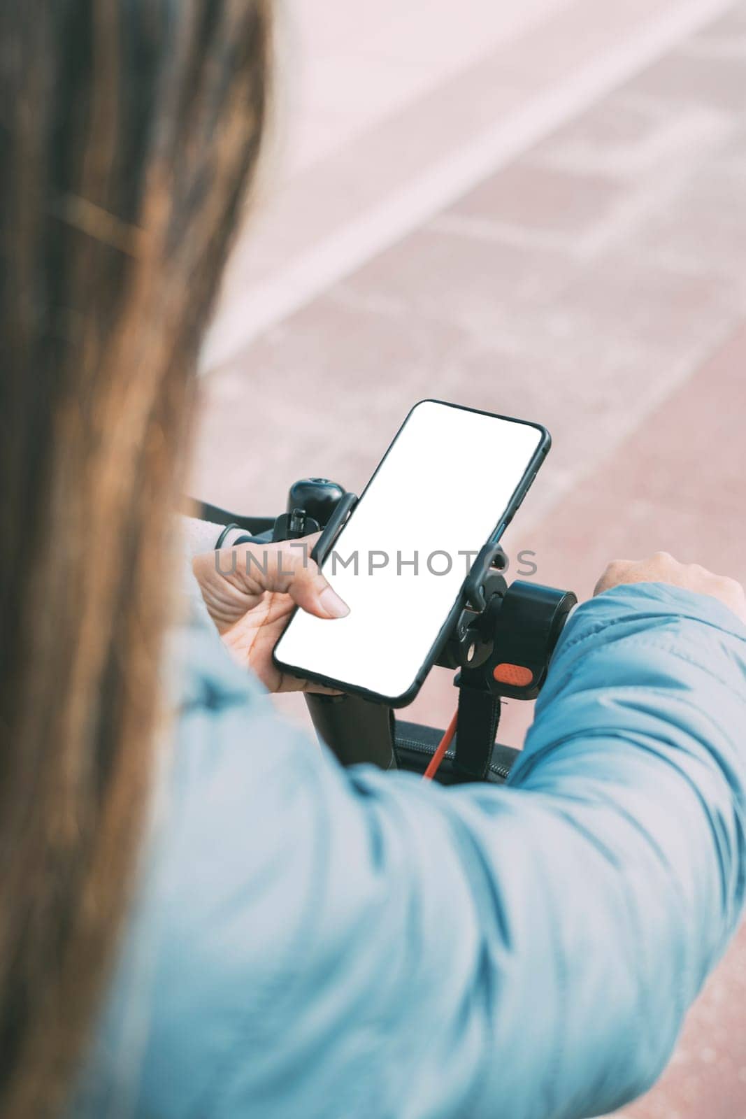 top rear view of middle-aged latina woman in her 40s 50s with long hair and blue jacket riding electric scooter using smart phone, with white screen, for apps.