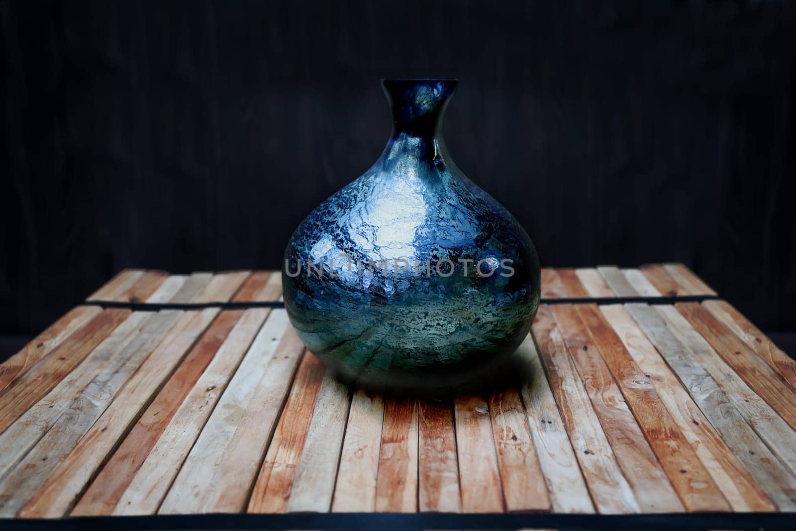 A still life with a blue vase on a stack of wooden slats, against a dark background.