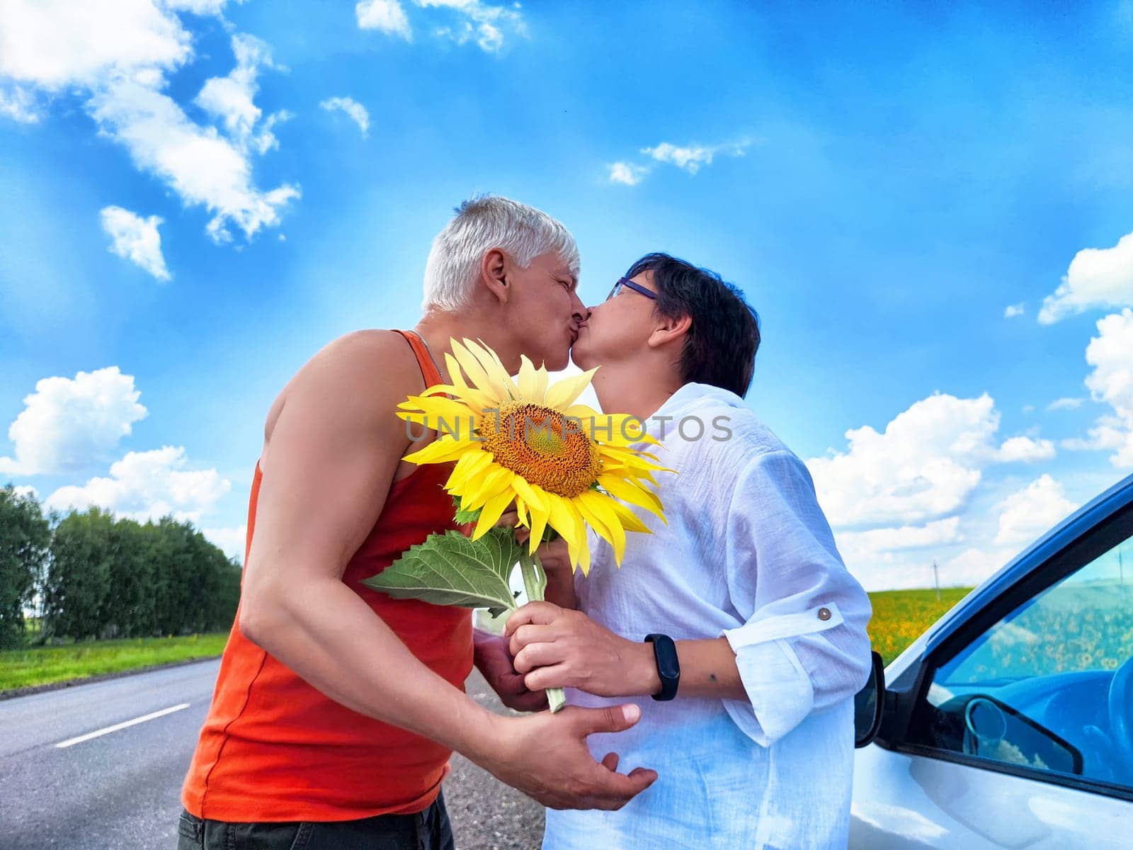 A middle-aged couple shares a sweet kiss by the roadside, surrounded by clear blue skies and fluffy clouds. The man holds a bright sunflower, symbolizing love and joy during their trip.