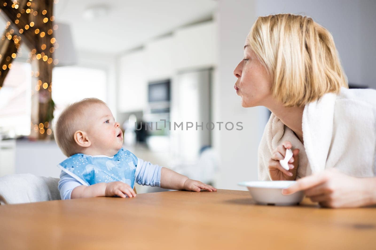 Mother motivating her baby boy infant child while spoon feeding him sitting in high chair at dinning table at home. Baby solid food introduction concept.