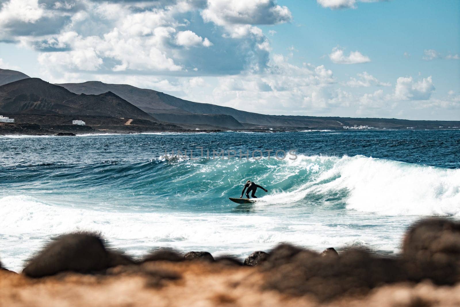 Wild rocky coastline of surf spot La Santa Lanzarote, Canary Islands, Spain. Surfer riding a big wave in rocky bay, volcano mountain in background
