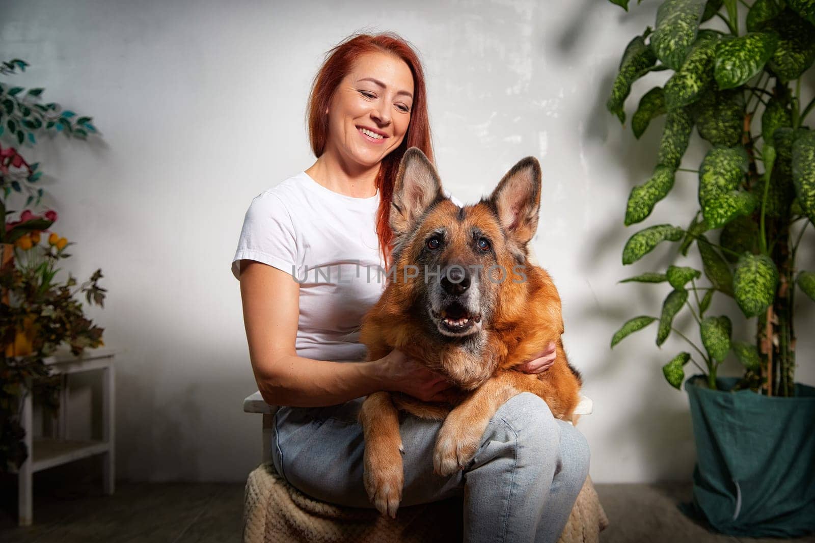 A woman sits comfortably with her German Shepherd dog, sharing a joyful moment in a well-lit room adorned with plants.