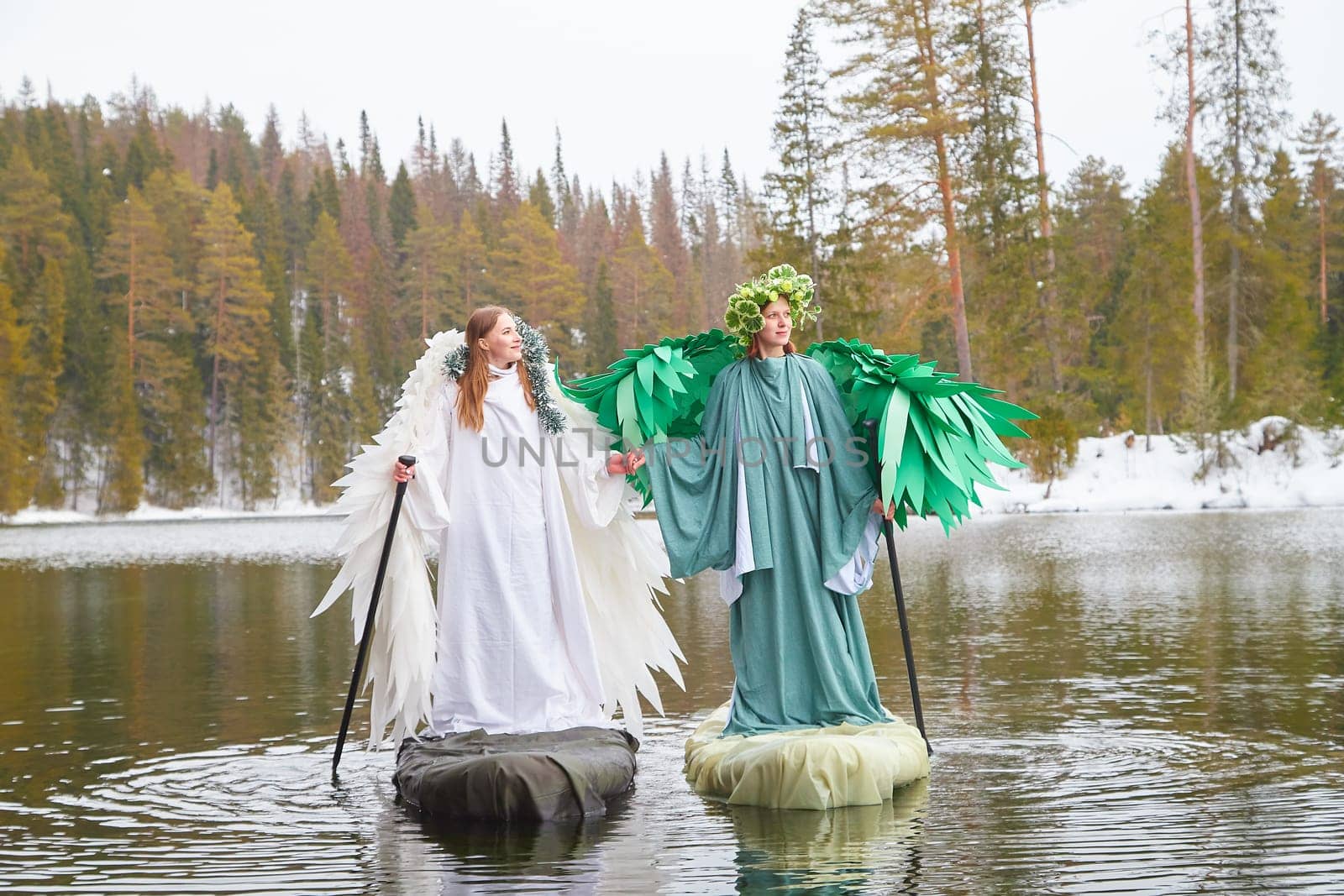 Two girls dressed in contrasting white and green outfits embody the essence of winter and spring beside a tranquil lake, capturing seasonal change.