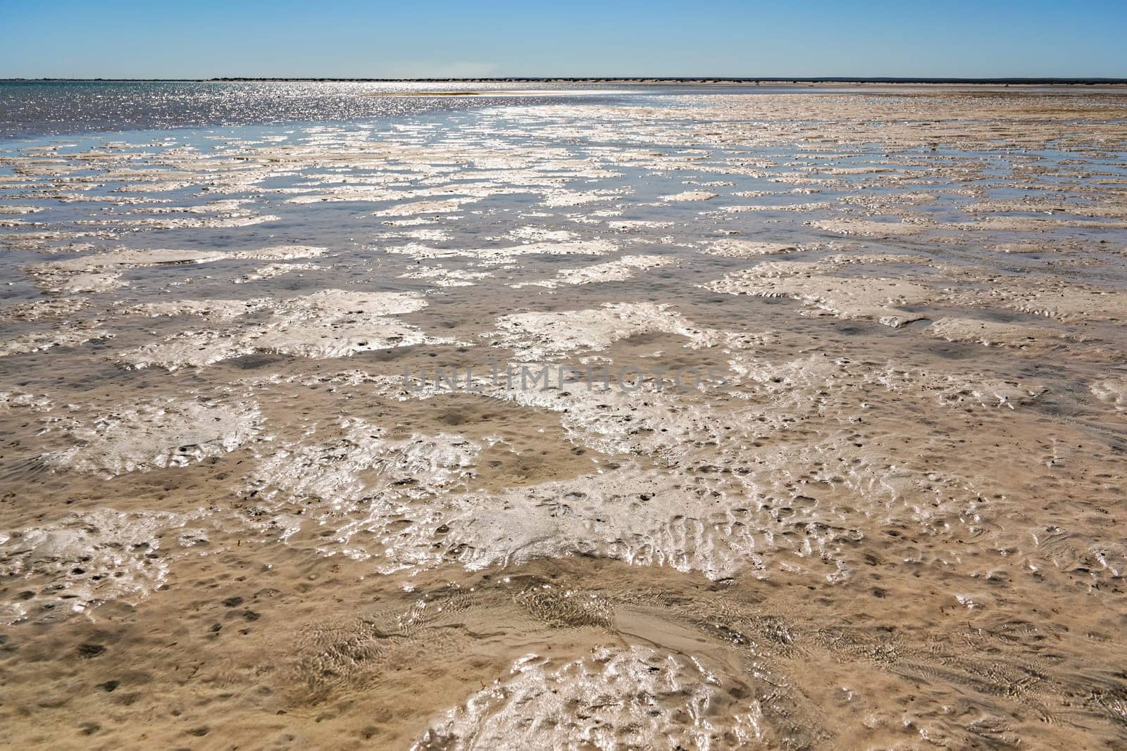 Sun shines to shallow sea during low tide, wet sand visible, sandy shore in distance.
