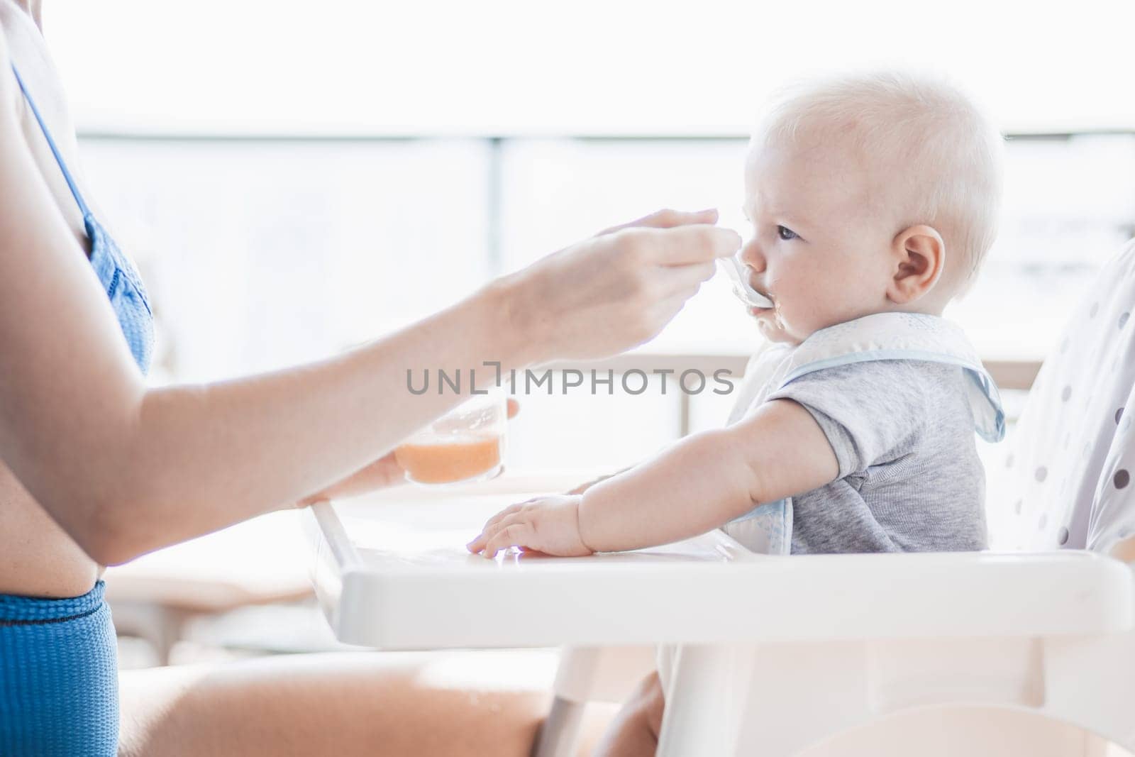 Mother spoon feeding her baby boy child in baby chair with fruit puree on a porch on summer vacations. Baby solid food introduction concept