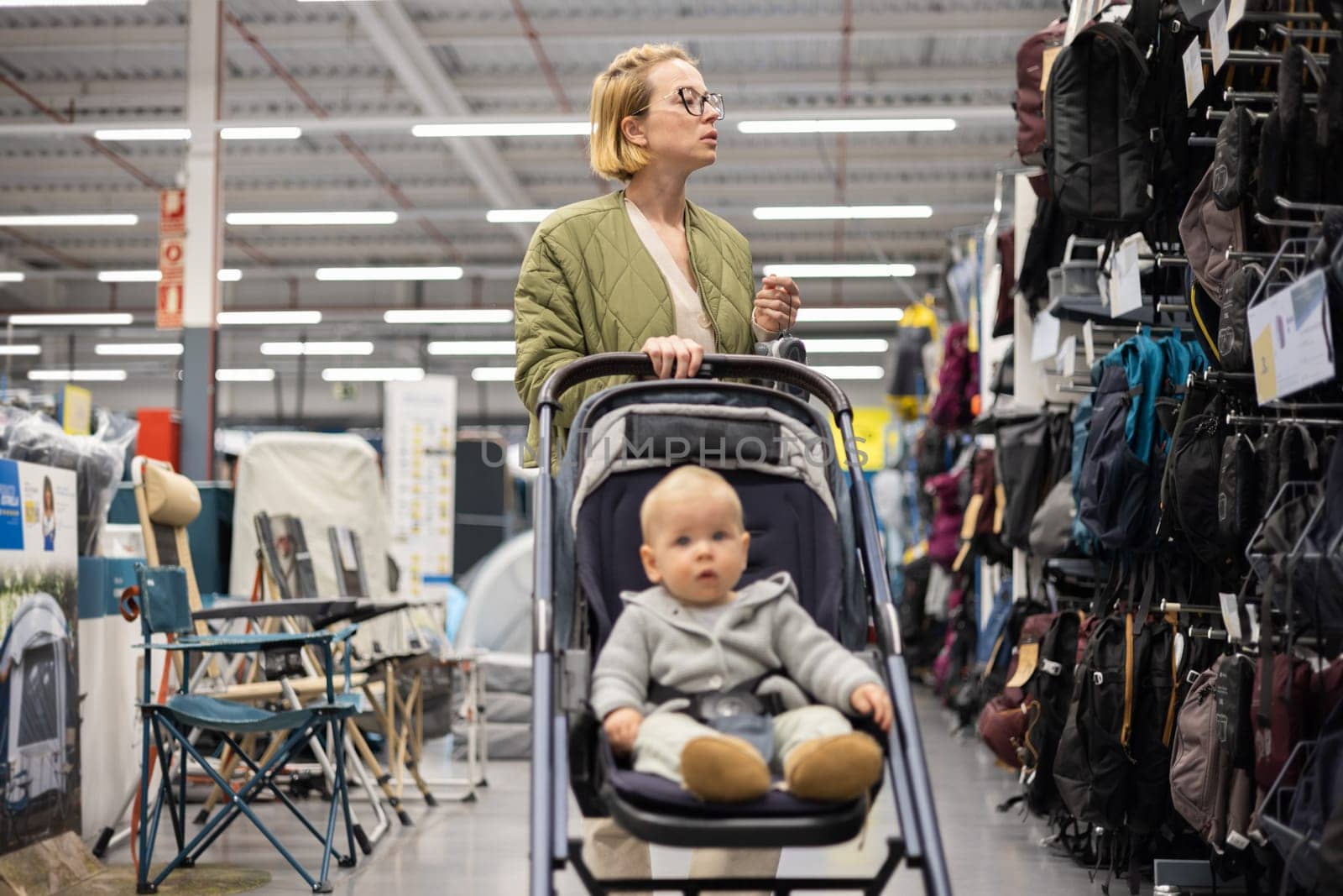 Casualy dressed mother choosing sporty shoes and clothes products in sports department of supermarket store with her infant baby boy child in stroller