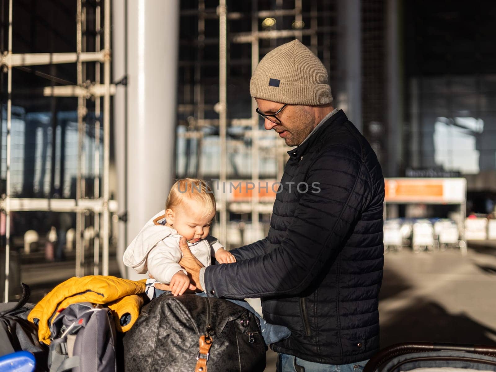 Fatherat comforting his tired infant baby boy child sitting on top of luggage cart in front of airport terminal station while traveling wih family