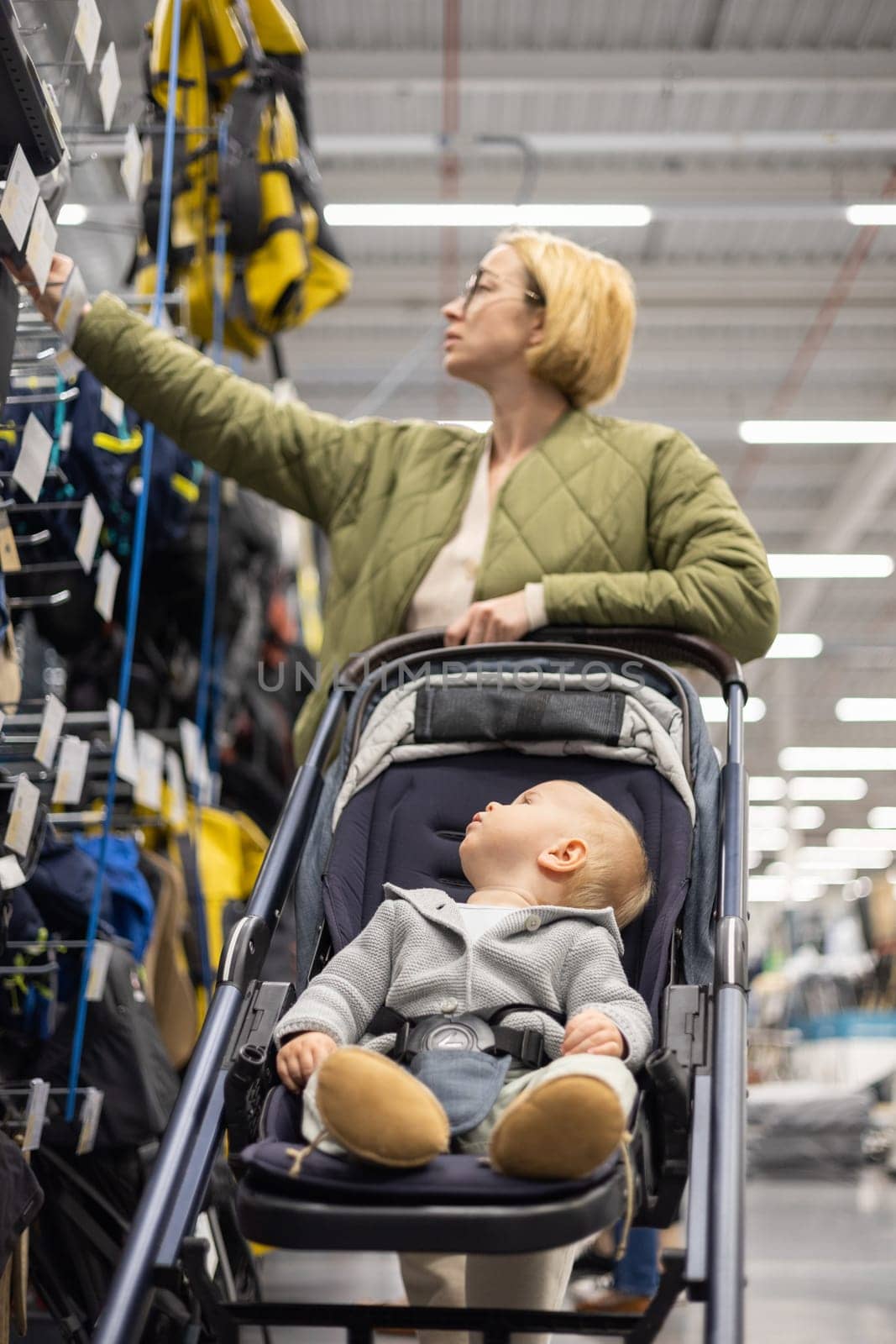 Casualy dressed mother choosing sporty shoes and clothes products in sports department of supermarket store with her infant baby boy child in stroller