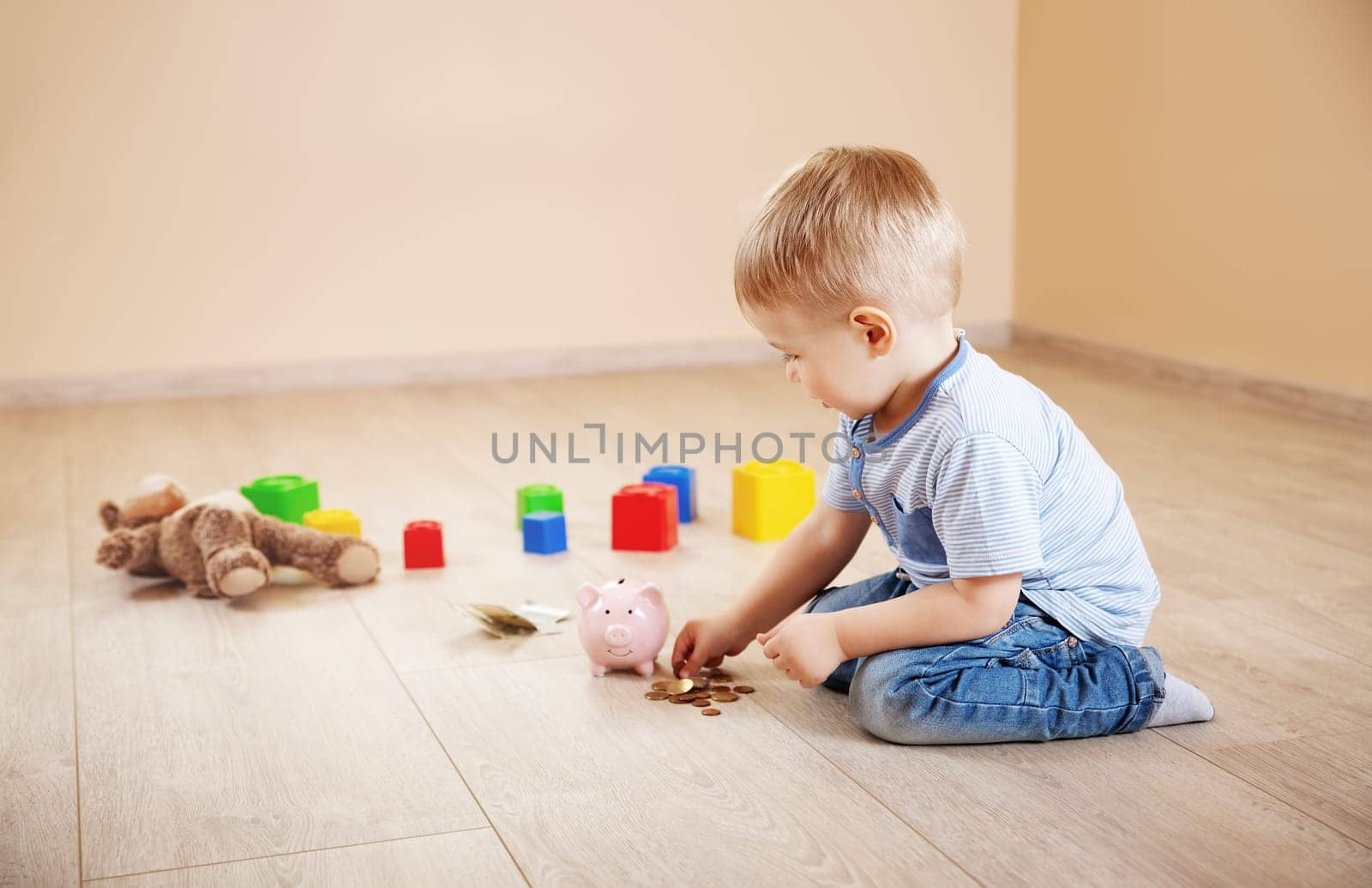 Cute boy sitting on the floor indoor and inserting euro coins in piggybank. Concept of the economy and family budget.
