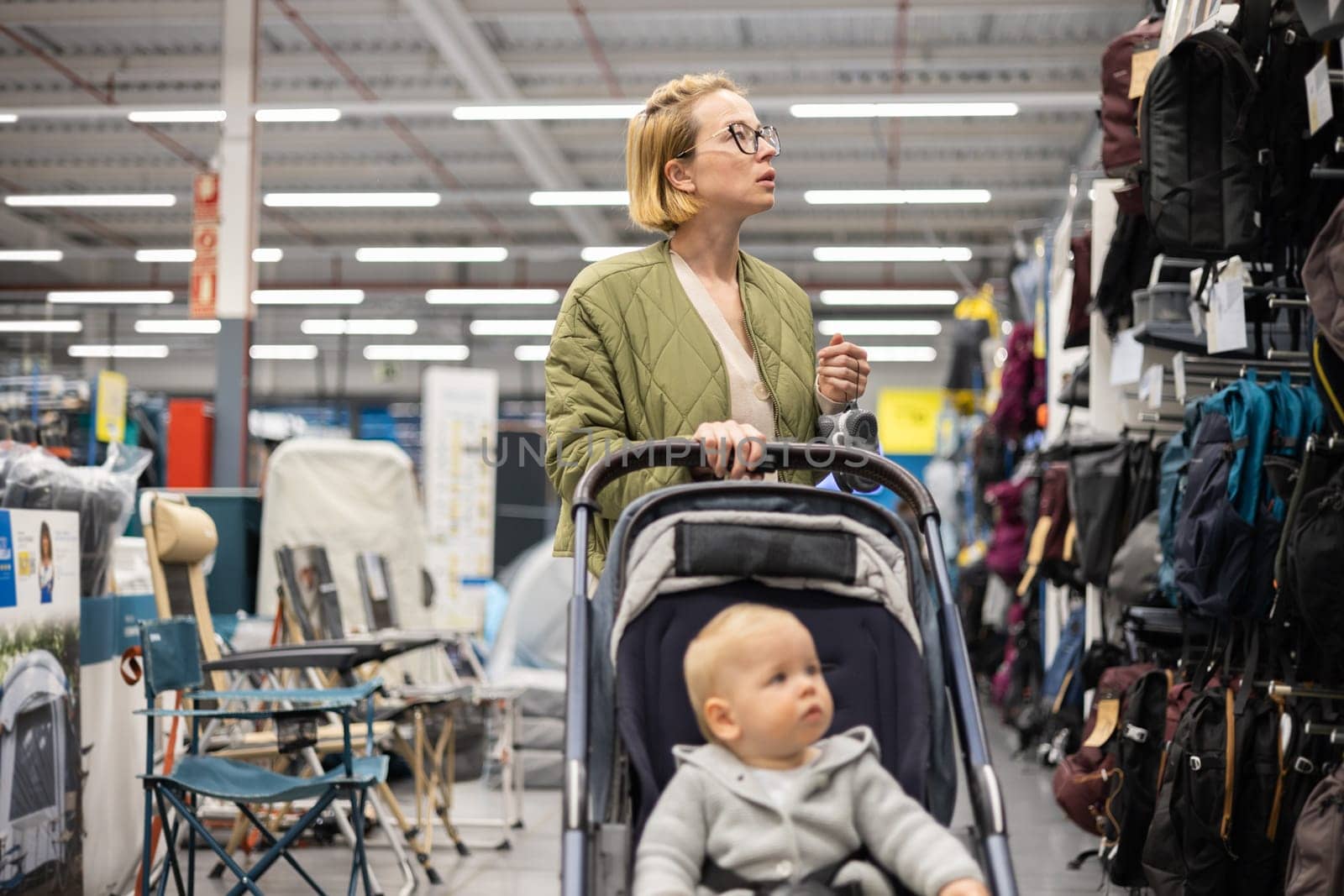 Casualy dressed mother choosing sporty shoes and clothes products in sports department of supermarket store with her infant baby boy child in stroller