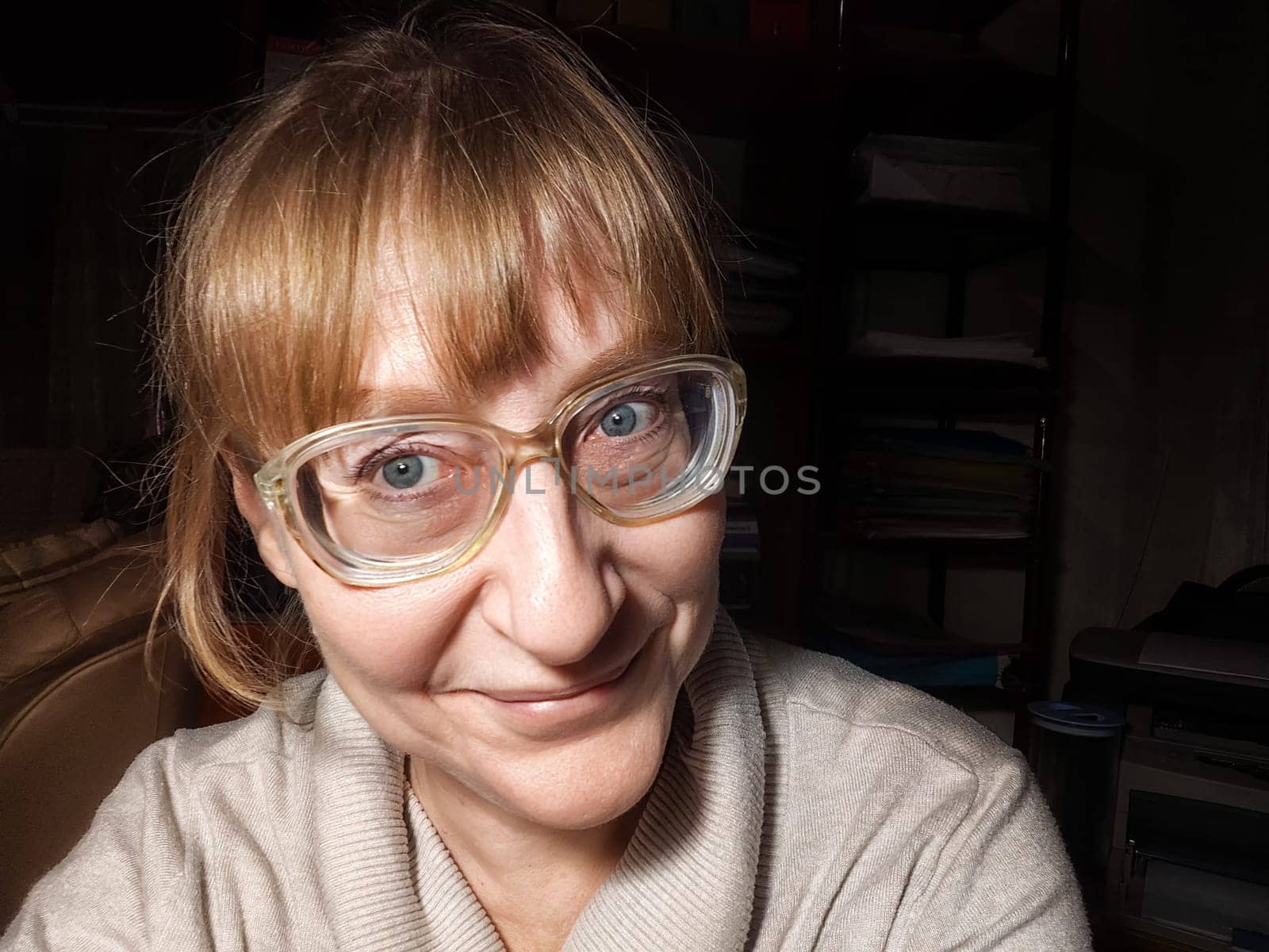A middle-aged woman with blonde hair smiles for a selfie, showcasing her playful side in a cozy indoor setting.