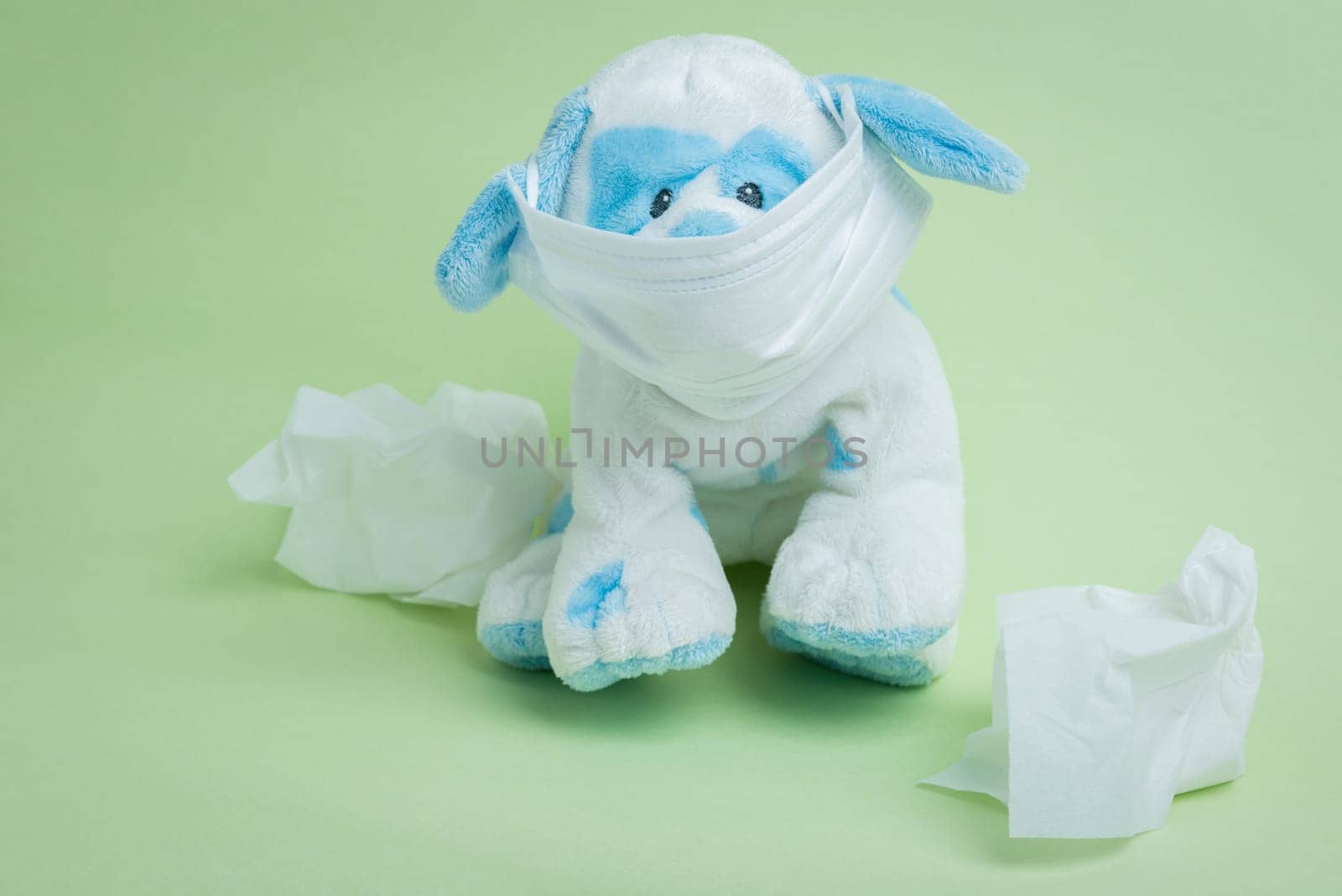 A white and blue stuffed dog wearing a surgical mask surrounded by used tissues representing the idiomatic phrase "sick as a dog."""