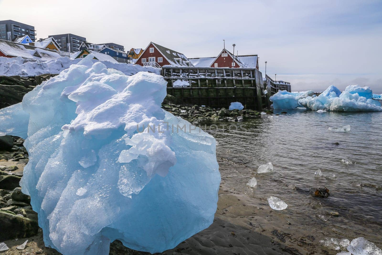 Big piece of blue ice laying among the stones on the shore with modern Inuit building on the hill at the fjord, Nuuk city, Greenland