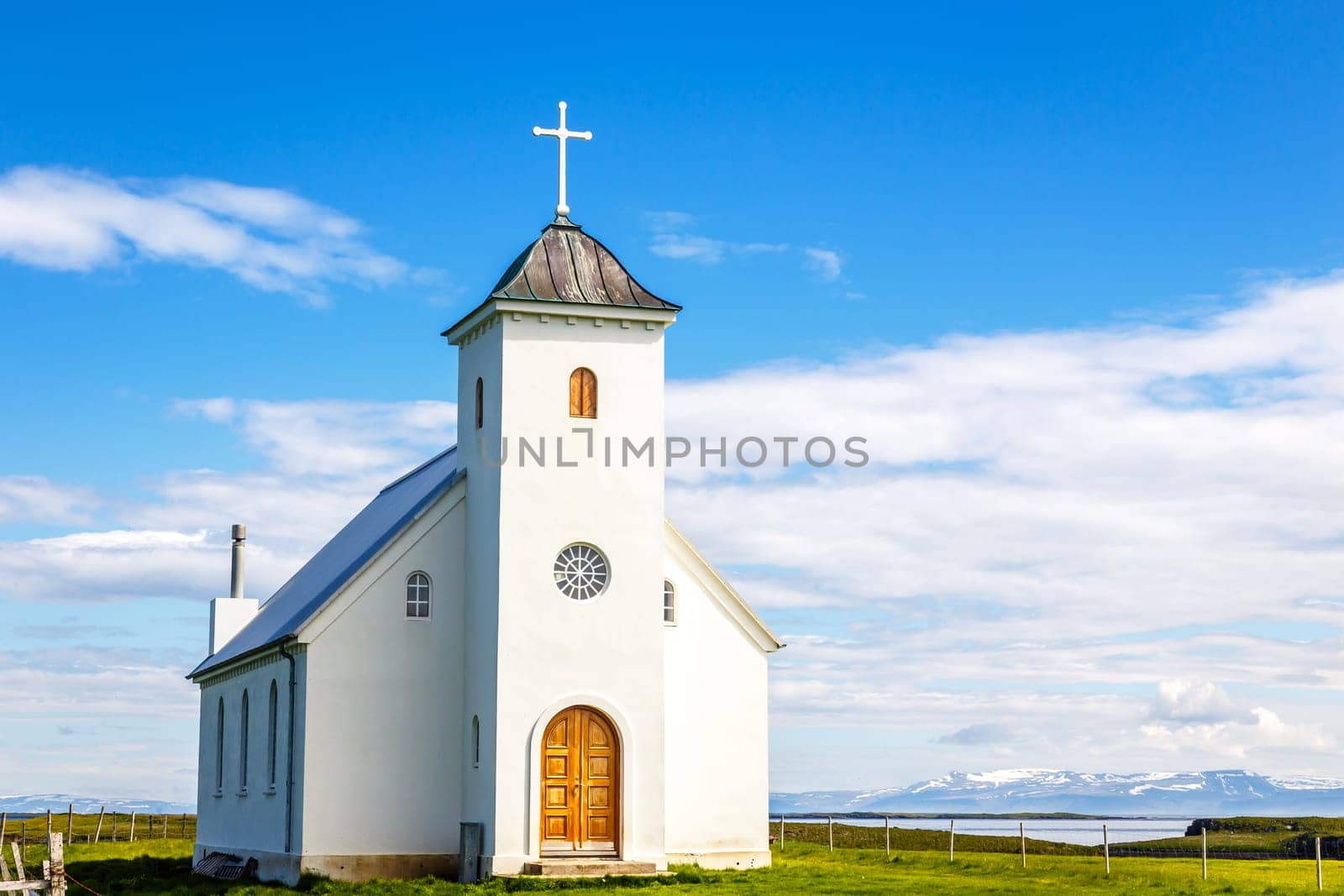 Flateyjarkirkja white lutheran church with meadow in foreground and sea  fjord with blue sky and mountains in the background, Flatey island, Iceland