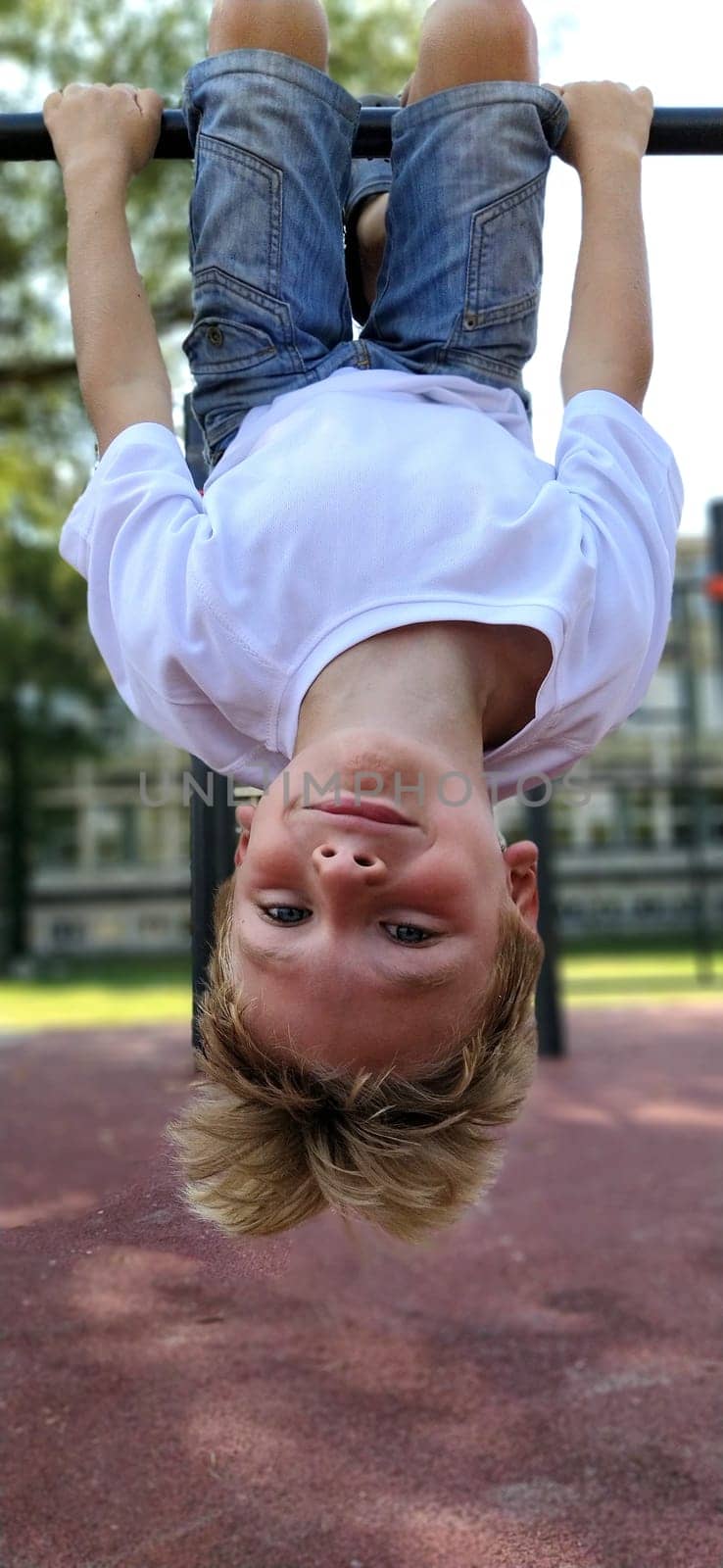 The boy hangs upside down on the sports ground. Exercises on sports equipment. Caucasian child 8 years old with blond hair. Sports and fun.
