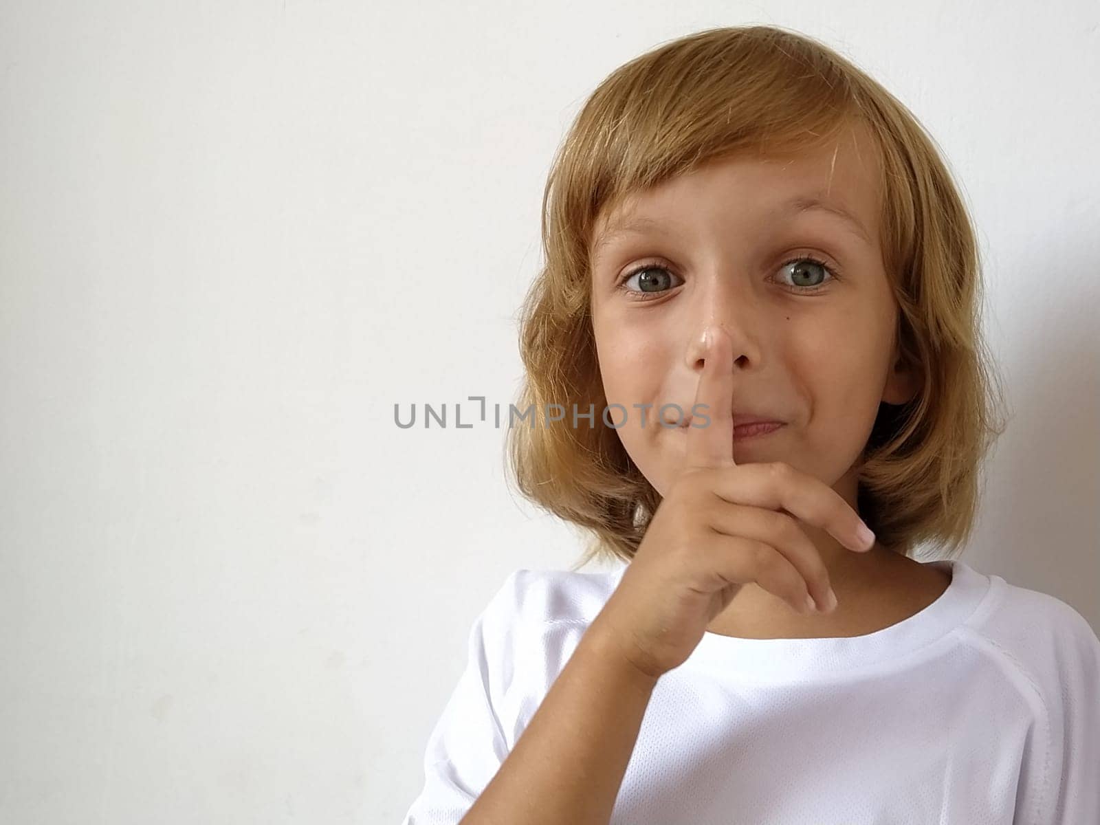 Silence gesture. The finger is near the mouth. The girl on a white background shows a gesture of keeping silence. Child of 7 years old with blond hair. Caucasian type. Copy space. Place for text.