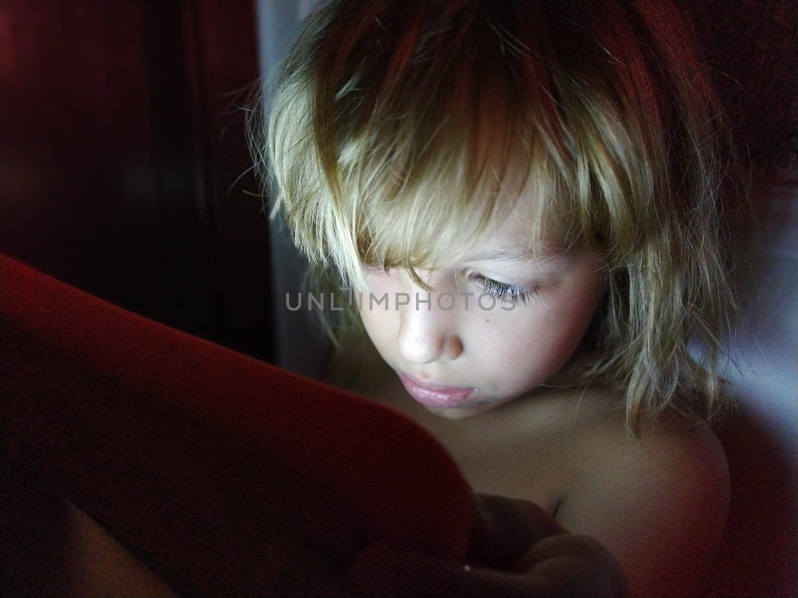 Cute little girl plays with a tablet computer. A child of 7 years old with a smiling face and blond hair looks into a gadget. Copy space. Girl in the room. Dark and light wall. At home in the evening.