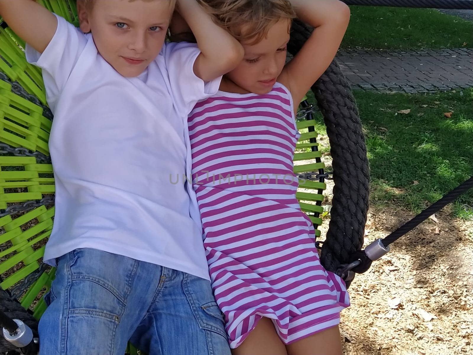 Children swing on a spider-web swing. Boy and girl, brother and sister in the summer on the playground. Swinging the swing. Boy 8 years old in a white Tshirt. Girl 6 years old in a striped pink dress