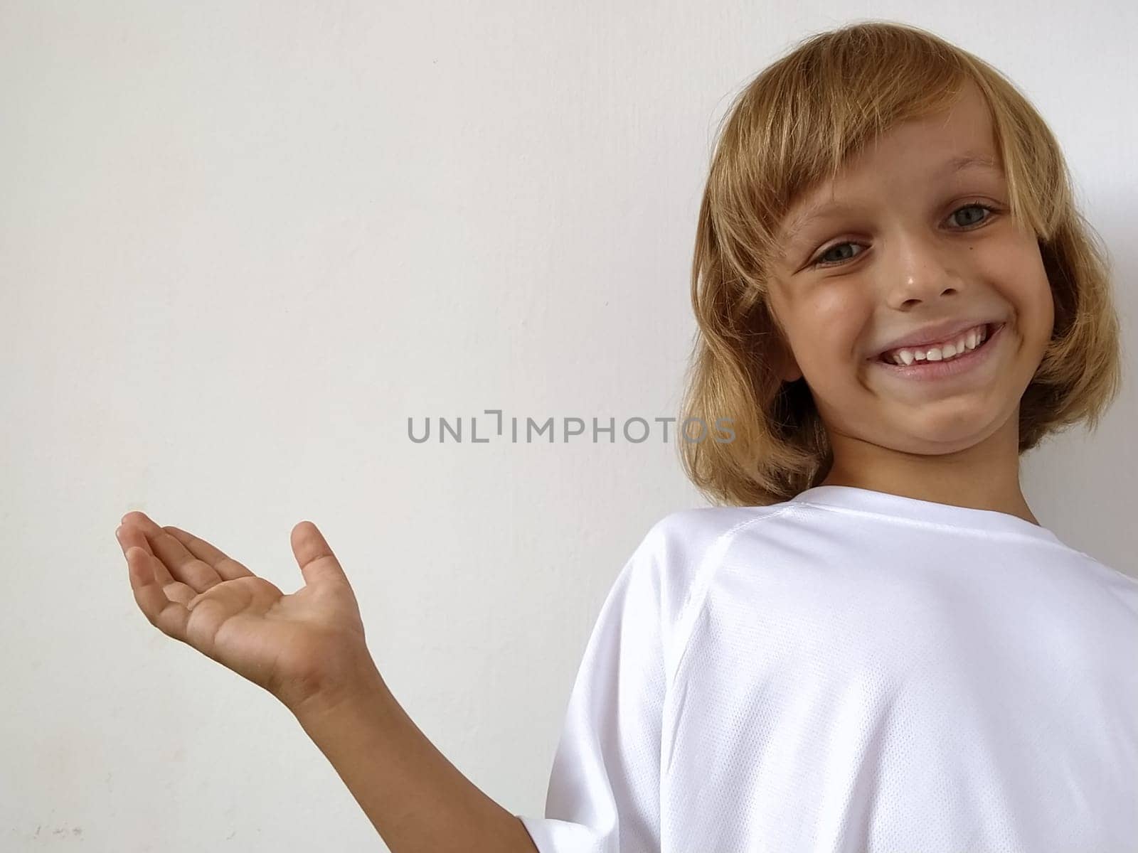 A little cute girl 6-7 years old with blond hair expresses emotional joy and a smile on a white background. A child with tanned skin. White T-shirt. Copy space. Girl with her hand points to something.