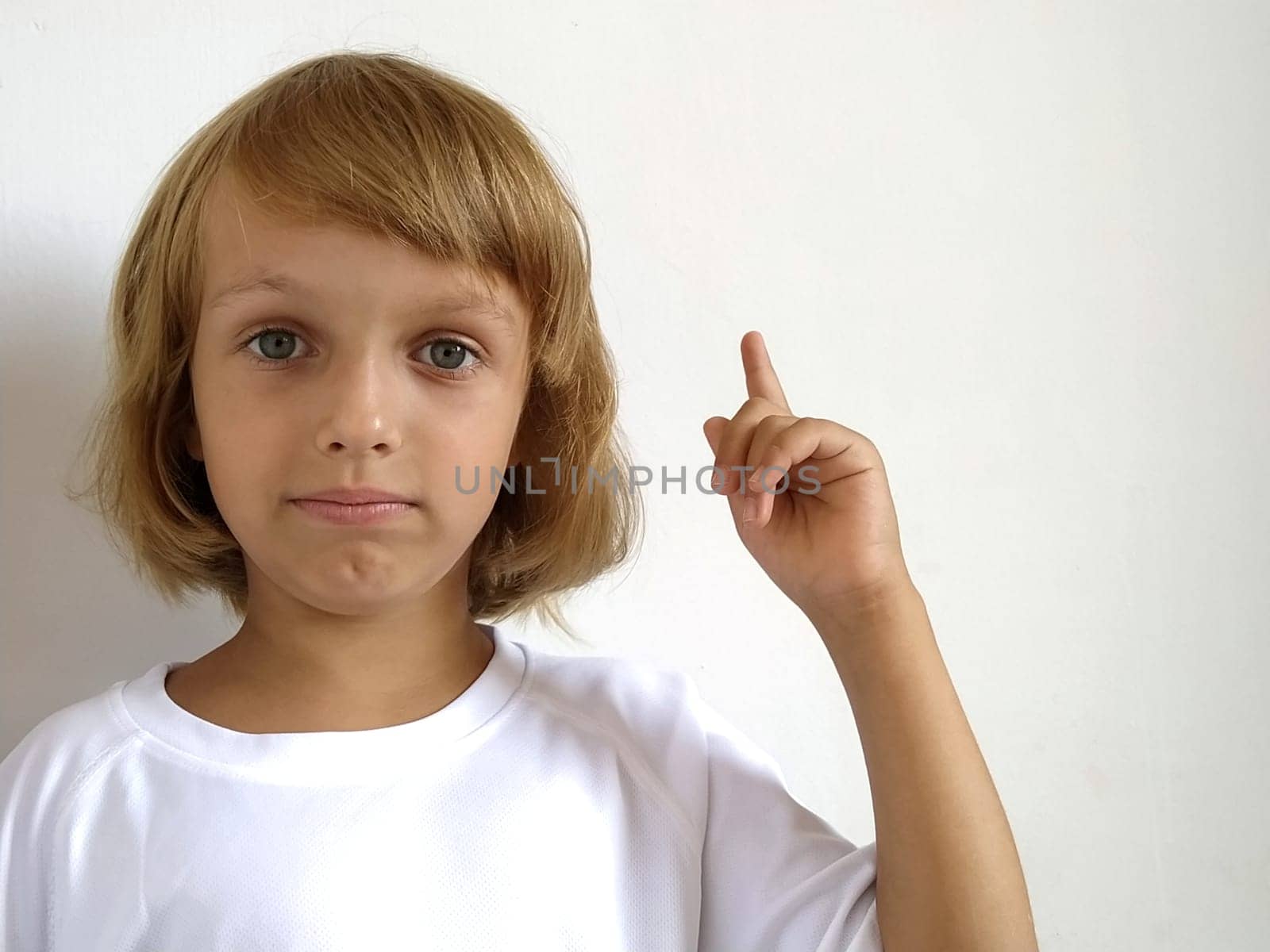 A little cute girl 6-7 years old with blond hair on a white background, pointing up. A child with tanned skin. White T-shirt. Copy space. Girl with her hand points to something. Bob hairstyle.