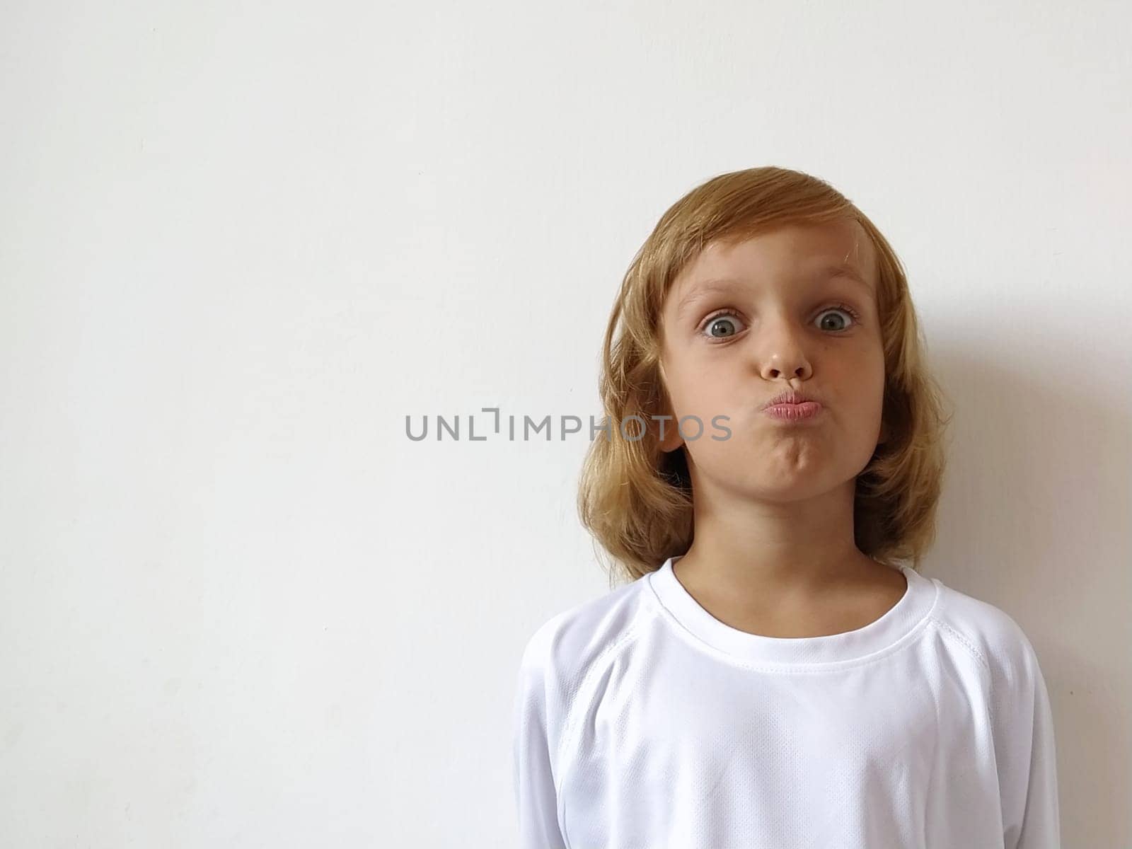 Little cute girl with blond hair on a white background. Tanned skin. White T-shirt. Copy space. The girl rolled her eyes and pursed lips. Facial expressions, surprise. Keeping a secret. Hairstyle Bob