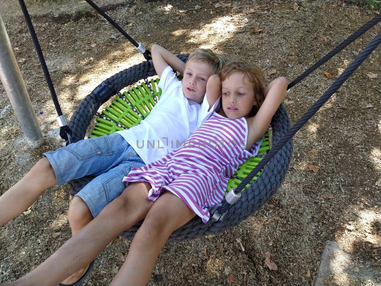 Children swing on a spider-web swing. Boy and girl, brother and sister in the summer on the playground. Swinging the swing. Boy 8 years old in a white shirt. Girl 6 years old in a striped pink dress.
