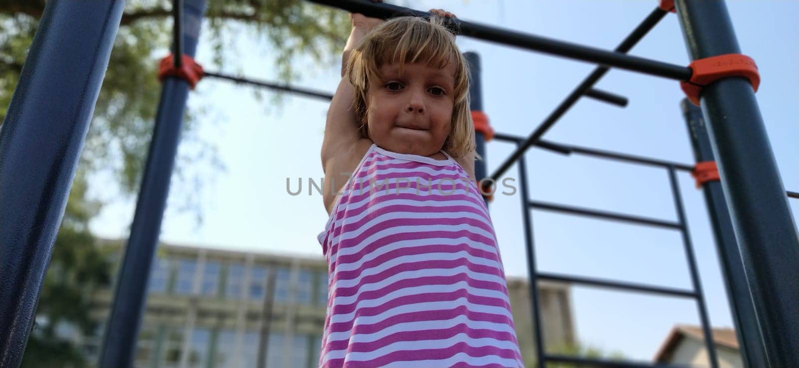A child in a striped dress hangs on a metal bar and tries to pull up on his arms. Sports or playground. Outdoor exercise.