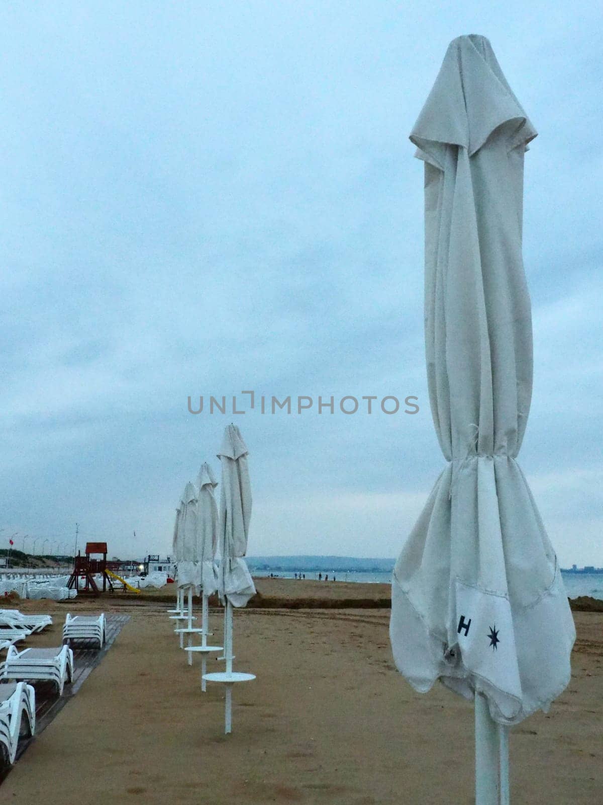 Wet sunbeds and umbrellas on the sea beach during heavy rain storm. Rolled up parasol on the beach against dark cloudy sky. Invasion of a typhoon, cyclone or storm on the sea or ocean. Beach season.