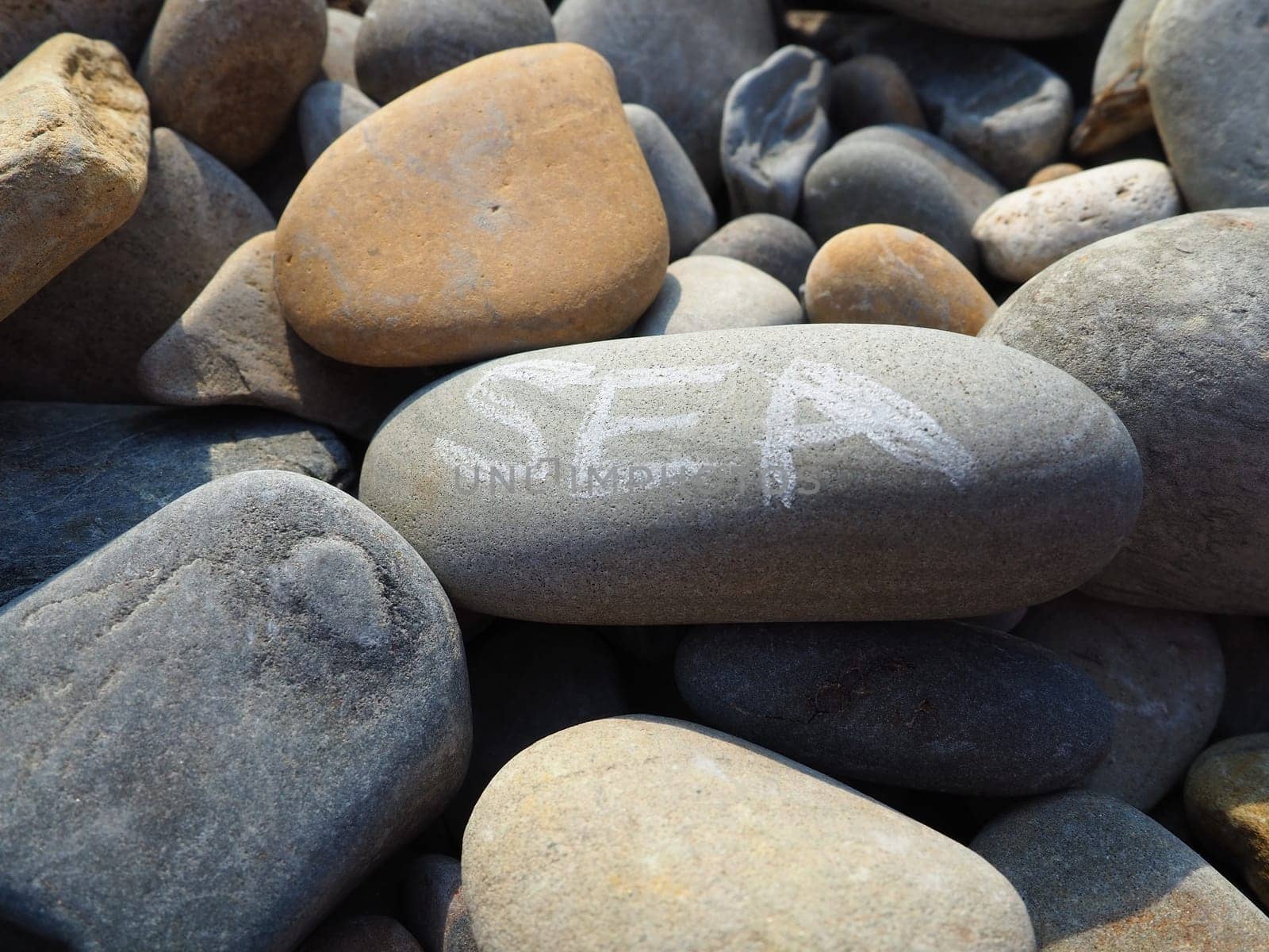 Oval stones, large smooth sea pebbles, illuminated by the summer sun. The word sea is written on the stone. Summer vacation in the resort, wellness, sea adventures and pleasures. Stone gray background