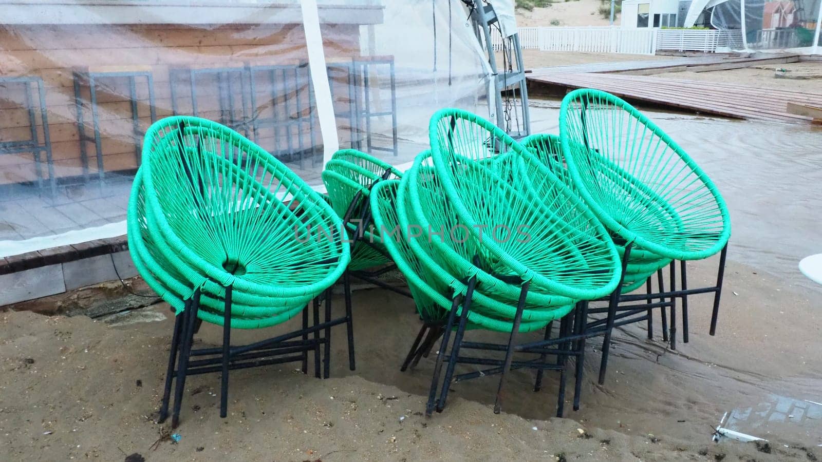 wicker green plastic chairs with black legs in a cafe on a sandy beach after a storm. The furniture is stacked on top of each other as the beach cafe is closed. End of the beach season at the hotel