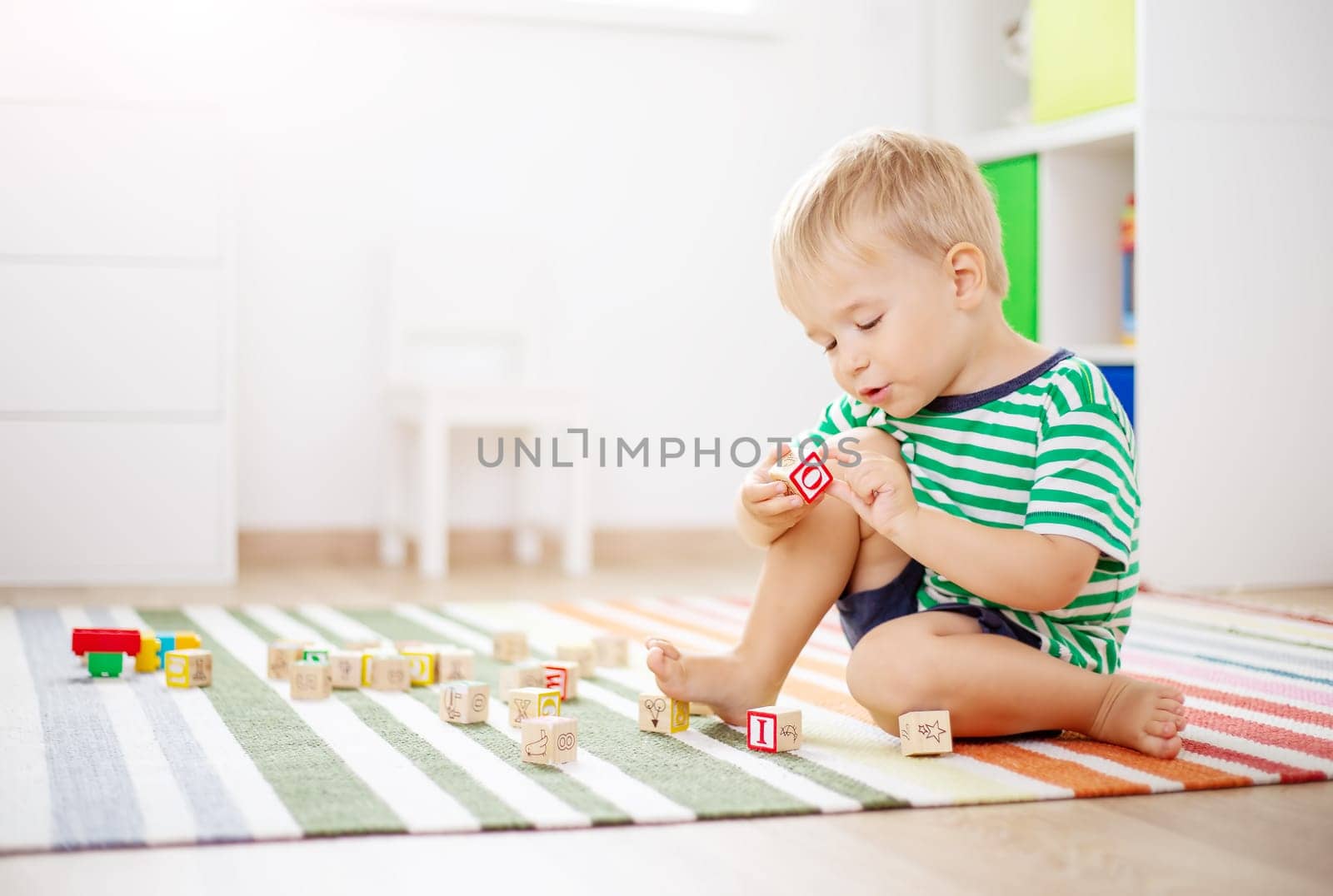 Cute boy sitting on the carpet indoor and playing with wooden cubes. Concept of preschool and kindergarden.