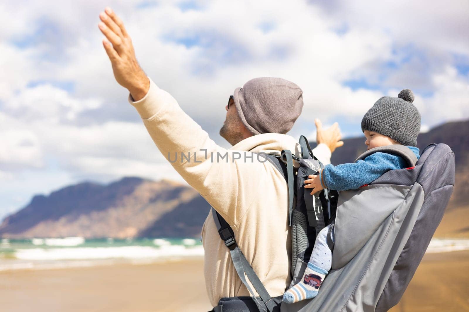 Young father rising hands to the sky while enjoying pure nature carrying his infant baby boy sun in backpack on windy sandy beach of Famara, Lanzarote island, Spain. Family travel concept