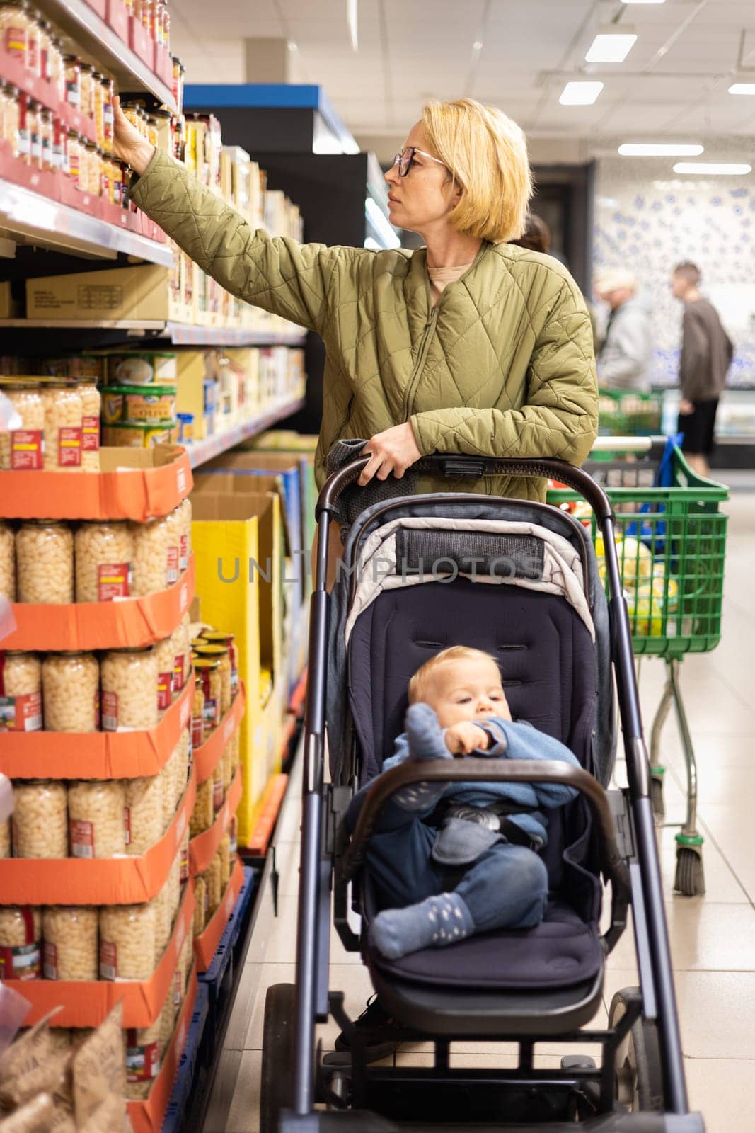 Casualy dressed mother choosing canned products in department of supermarket grocery store with her infant baby boy child in stroller
