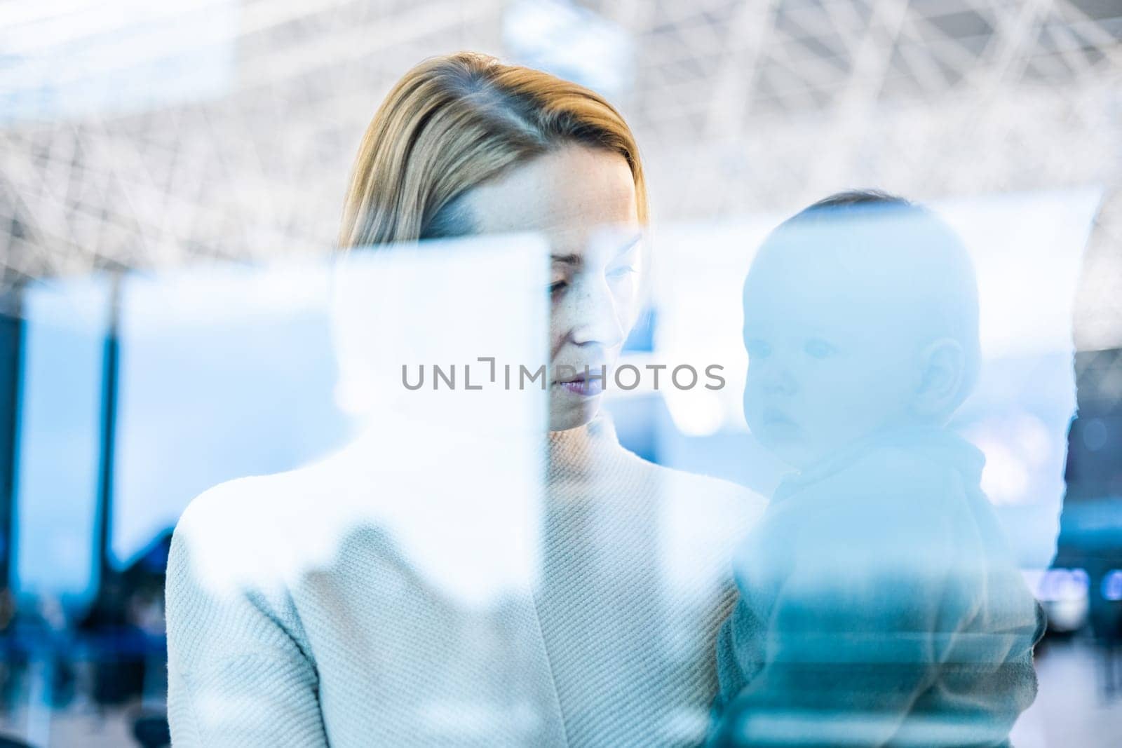 Thoughtful young mother looking trough window holding his infant baby boy child while waiting to board an airplane at airport terminal departure gates. Travel with baby concept