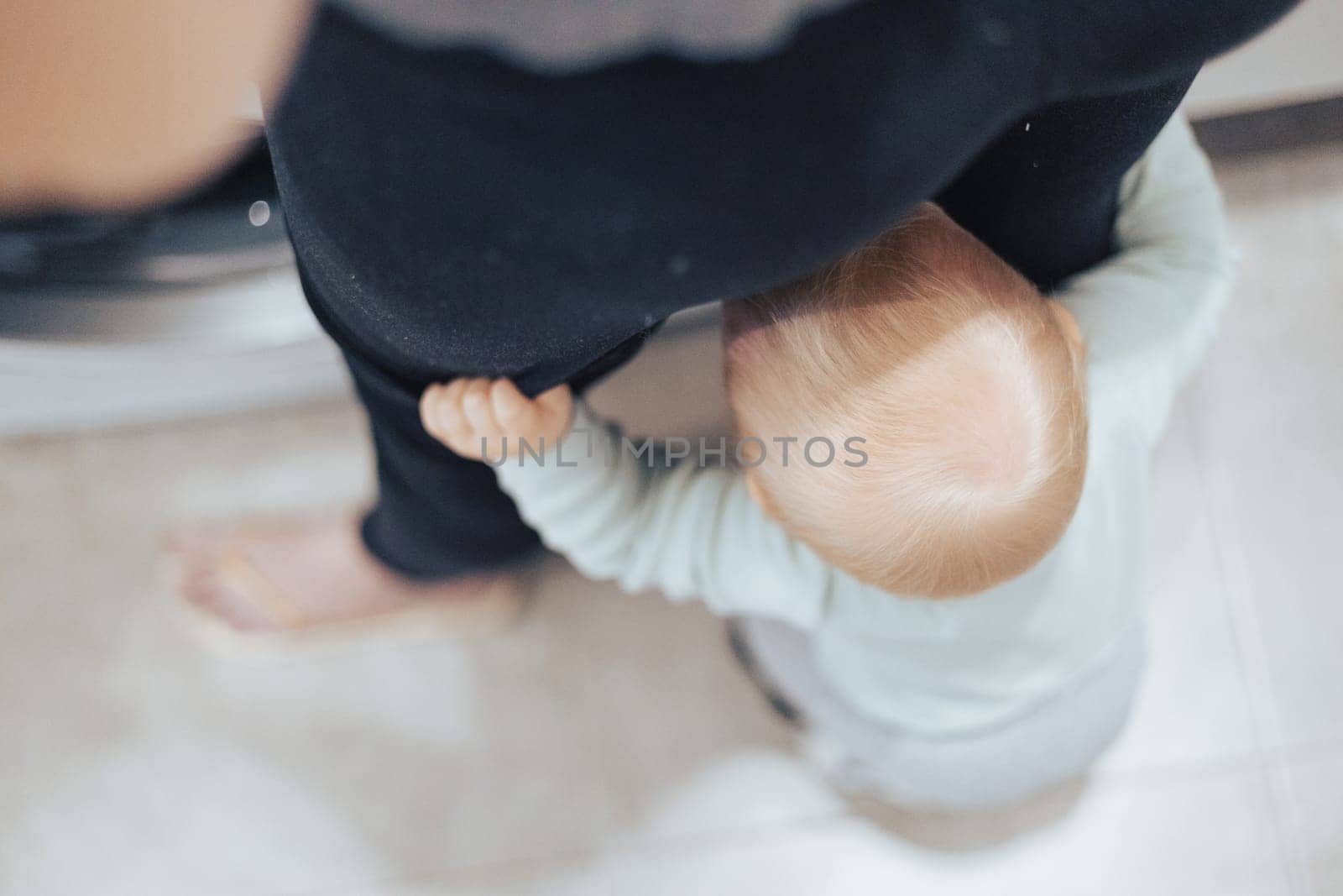 Top down view of little infant baby boy hiding between mothers legs demanding her attention while she is multitasking, trying to do some household chores in kitchen at home. Mother on maternity leave